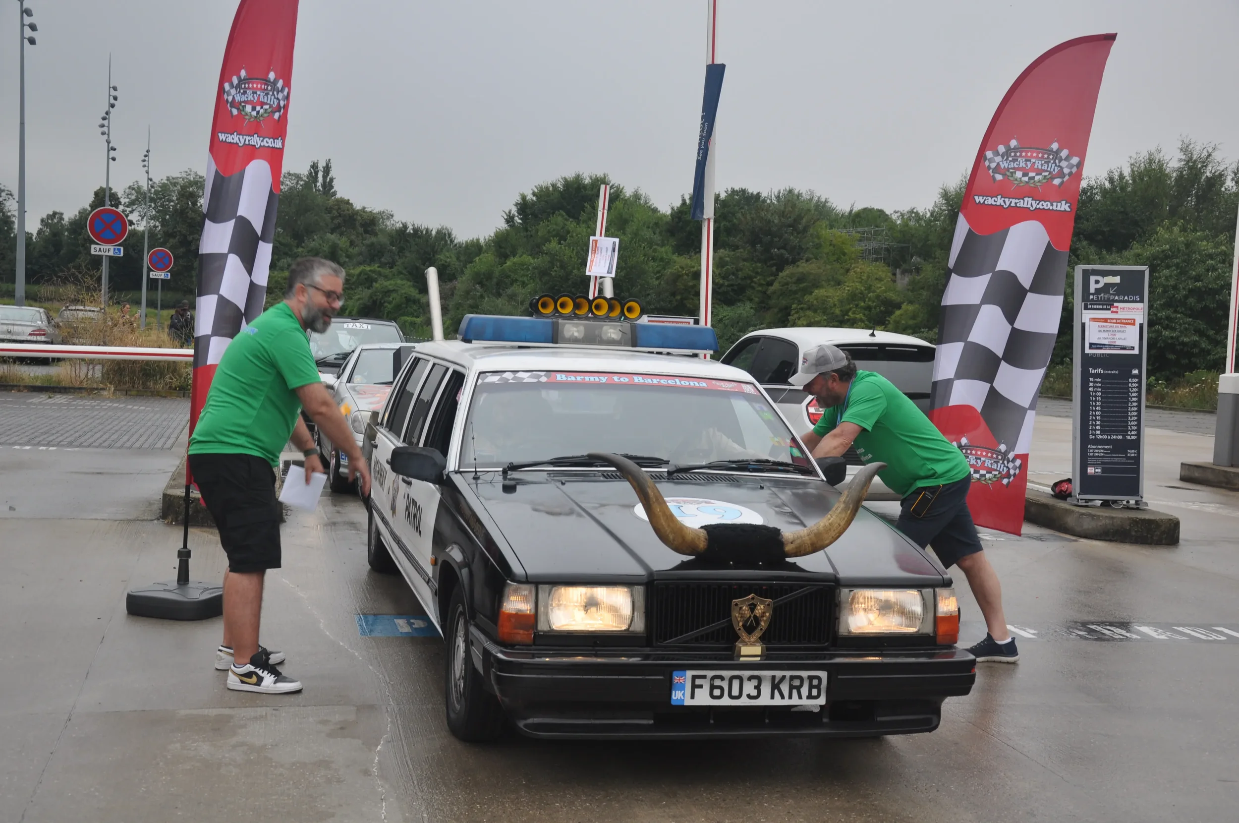 A black car with large horns mounted on the hood is parked in a lot with two men in green shirts cleaning or working on it. Flag banners with checkered patterns and the logo 'Wacky Rally' are on either side. The car has a police light bar on the roof