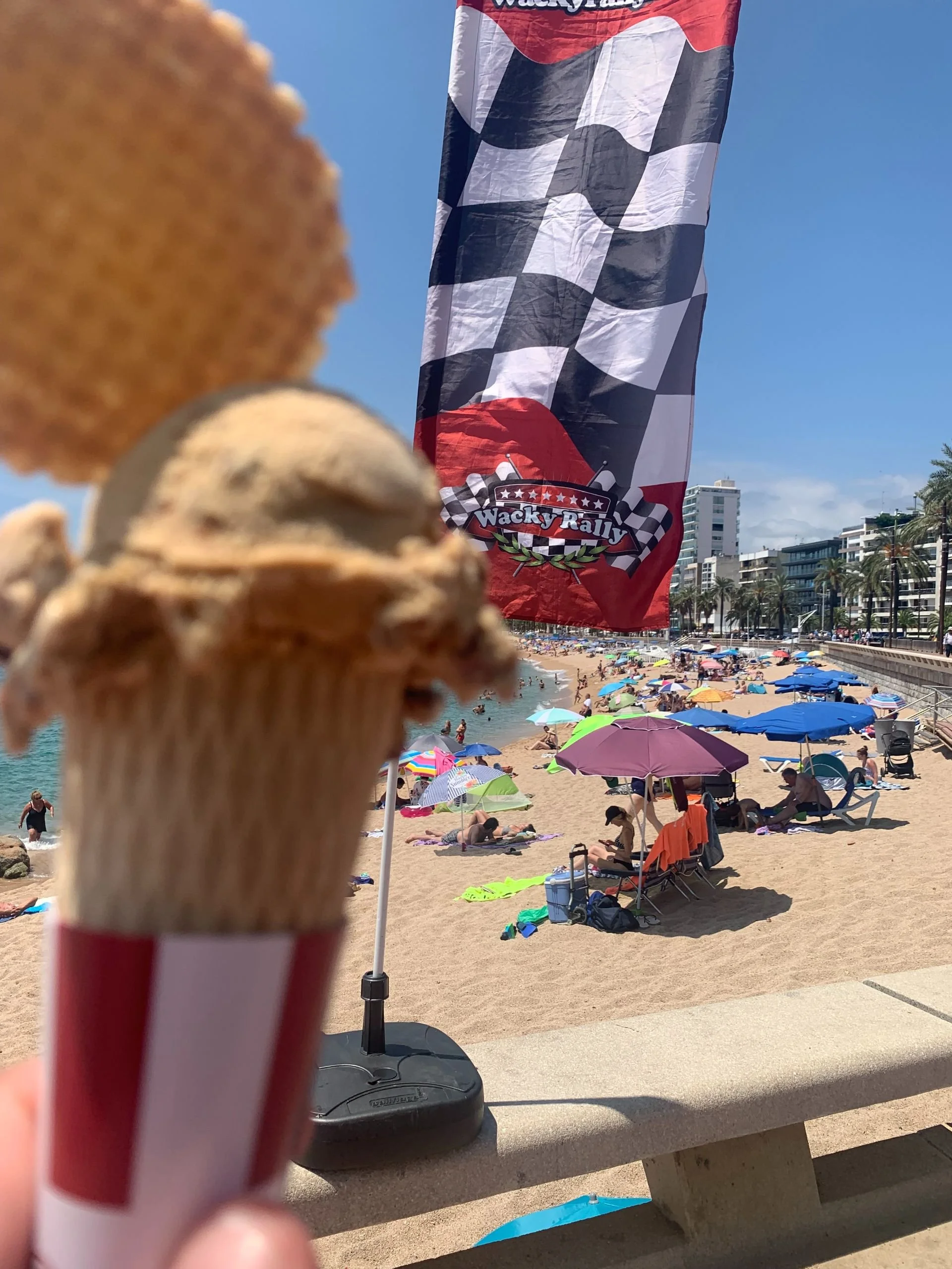 Close-up of a partially eaten vanilla ice cream cone with some cookies or biscuit on top, held up against a crowded beach scene with umbrellas and a red flag that says 'Wacky Rally'.