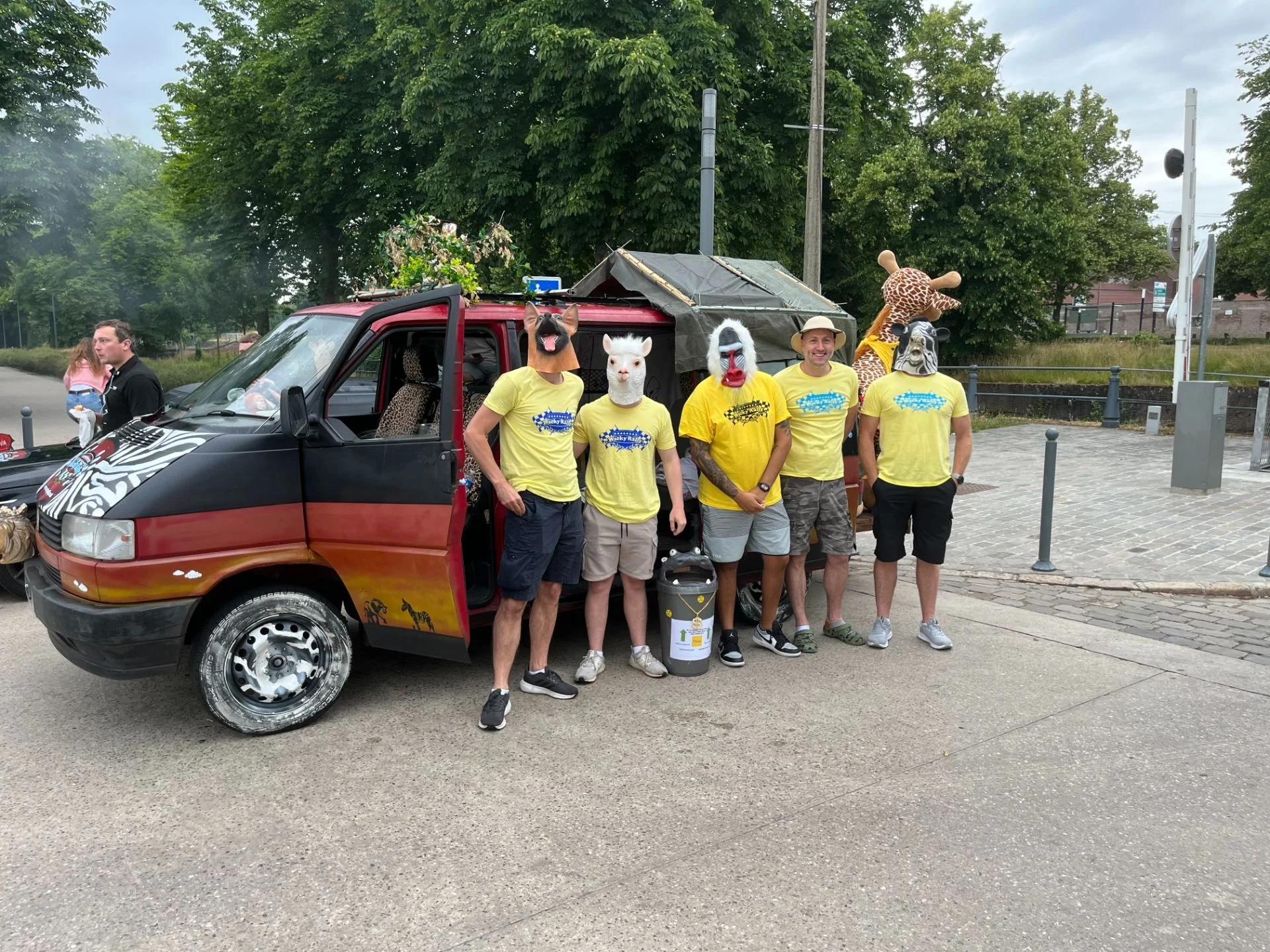 Group of five people wearing animal masks and yellow t-shirts, standing in front of a painted vehicle with a stuffed animal giraffe on top, outdoors on a cloudy day.