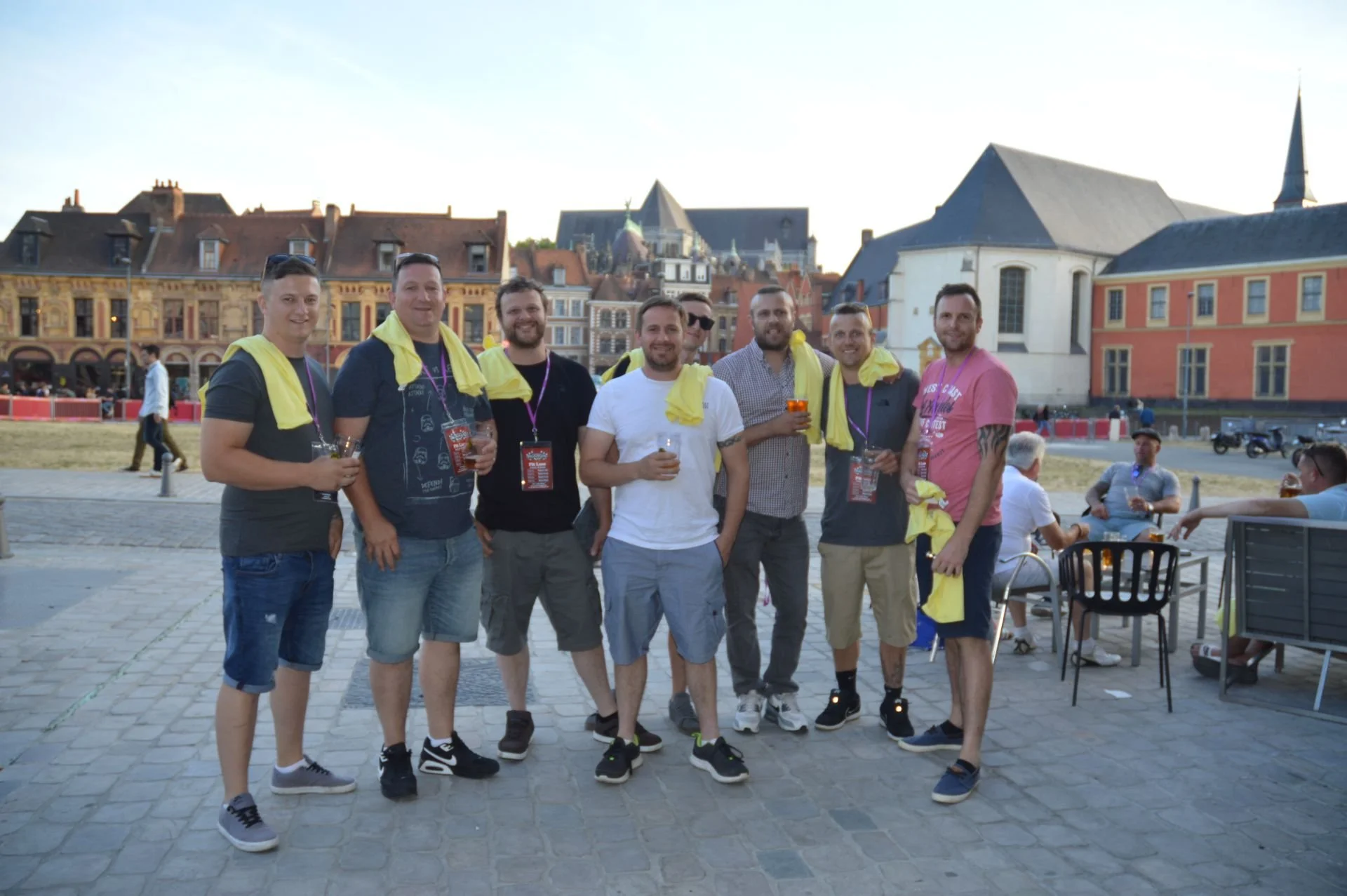 A group of nine smiling men standing outdoors on a cobblestone pavement, holding drinks, with some wearing yellow scarves around their necks, in front of historic European-style buildings with colorful facades.