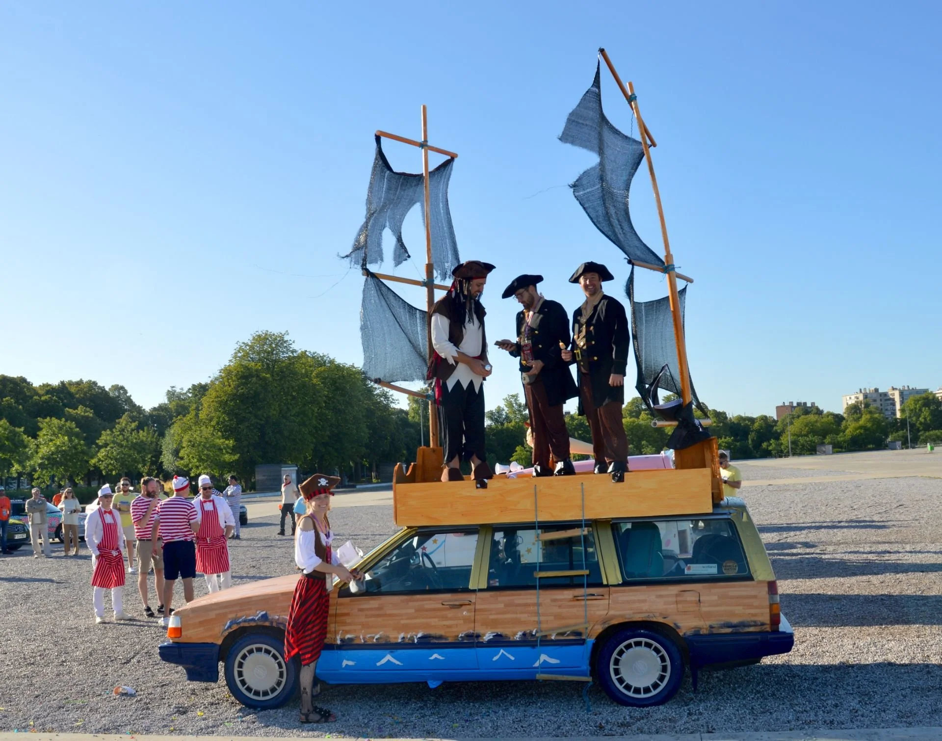 People dressed as pirates on top of a decorated vehicle resembling a pirate ship with sails, while others in striped outfits stand nearby in a large open area with trees and buildings in the background.