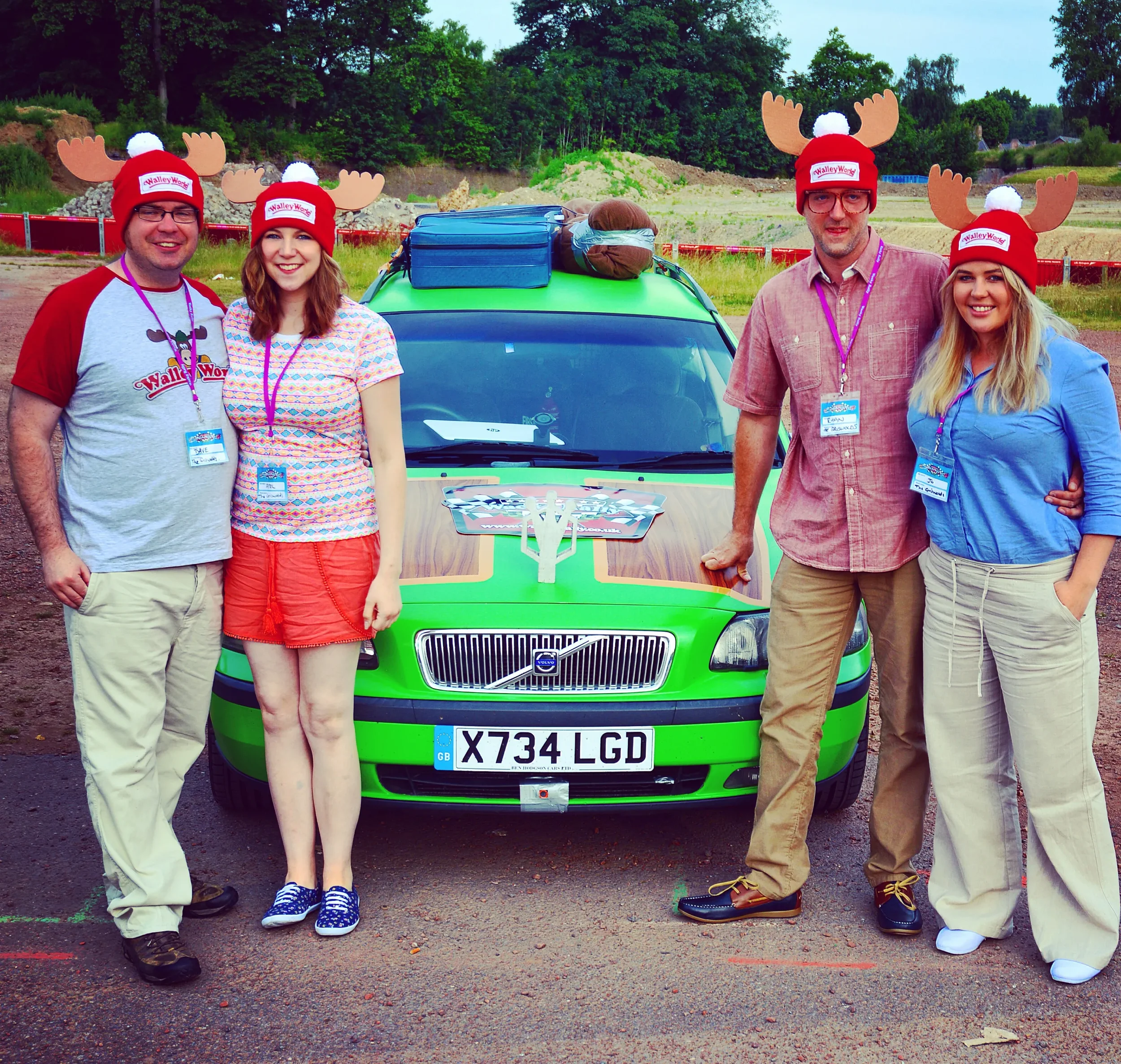 Four people standing in front of a green Volvo race car wearing Christmas-themed reindeer antler hats. They are outdoors on a dirt area with trees and construction in the background. Two men and two women, all smiling, are wearing casual clothes and 