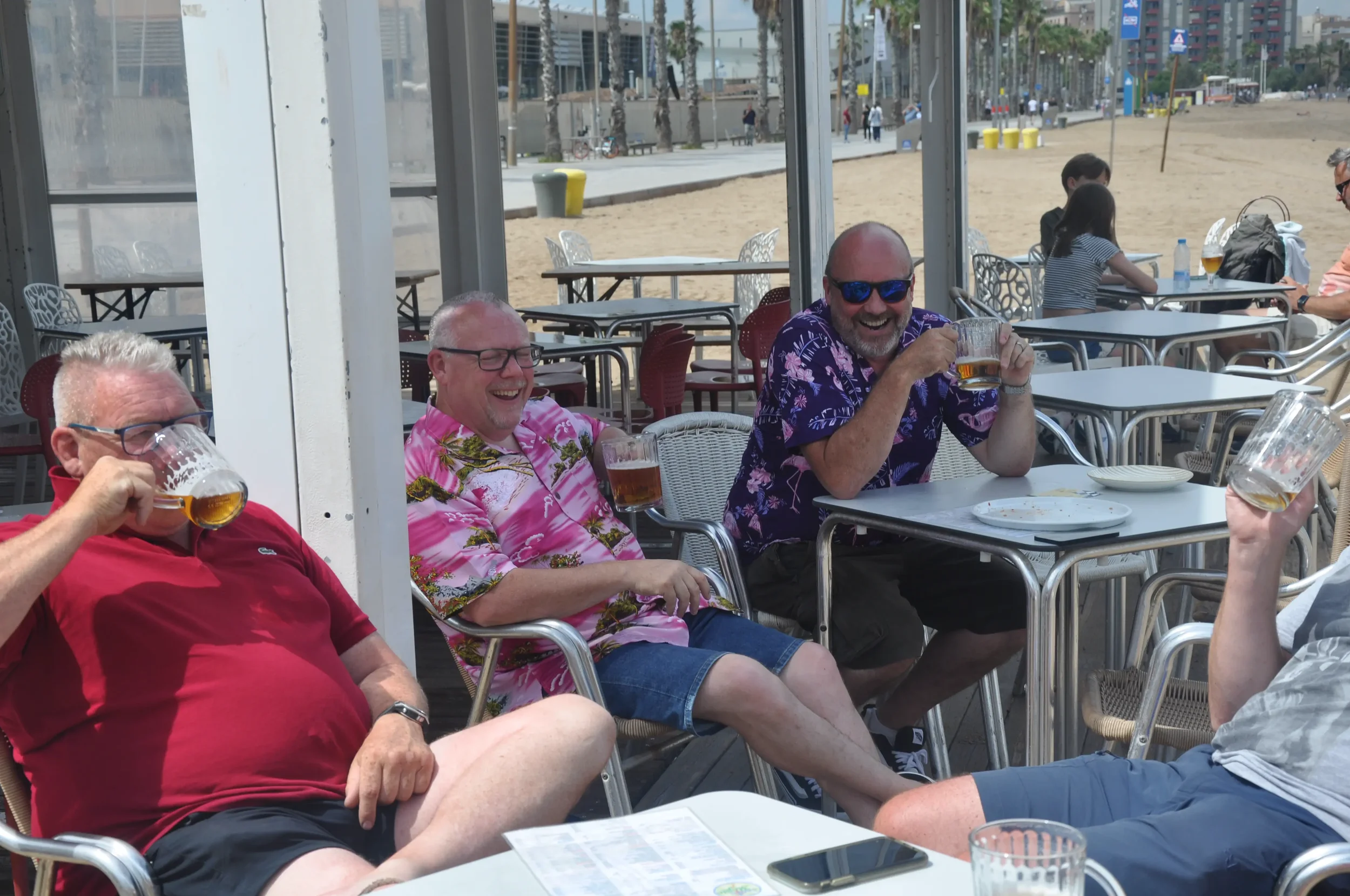 Four men are sitting at a table on an outdoor patio by the beach, drinking beer and laughing.