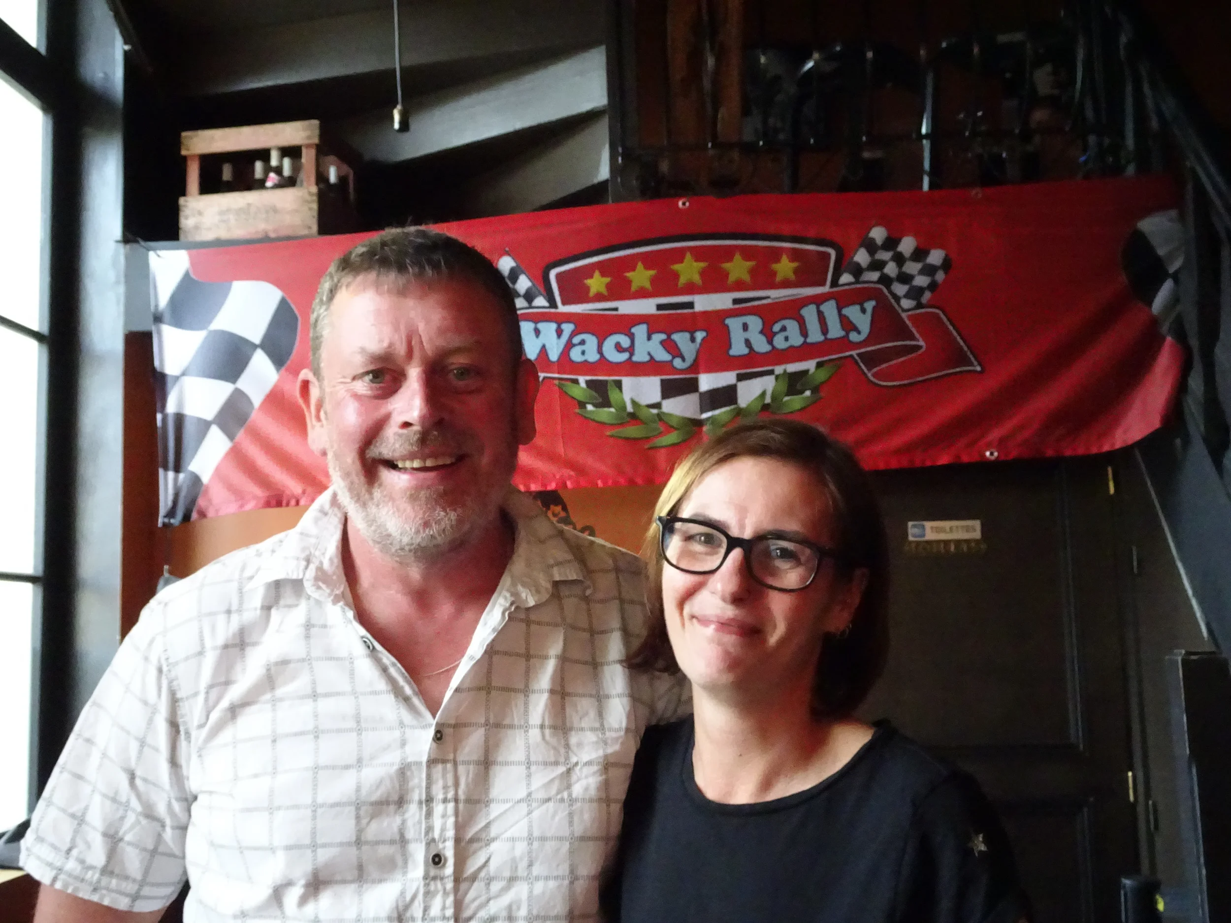 A man and a woman are standing close together inside a building, smiling at the camera. Behind them, a red banner with checkered flags and the words 'Wacky Rally' is displayed.