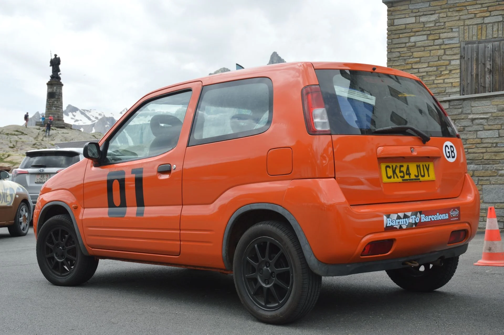 Orange compact car with racing numbers 01 on the side, parked near a stone building, with mountains in the background