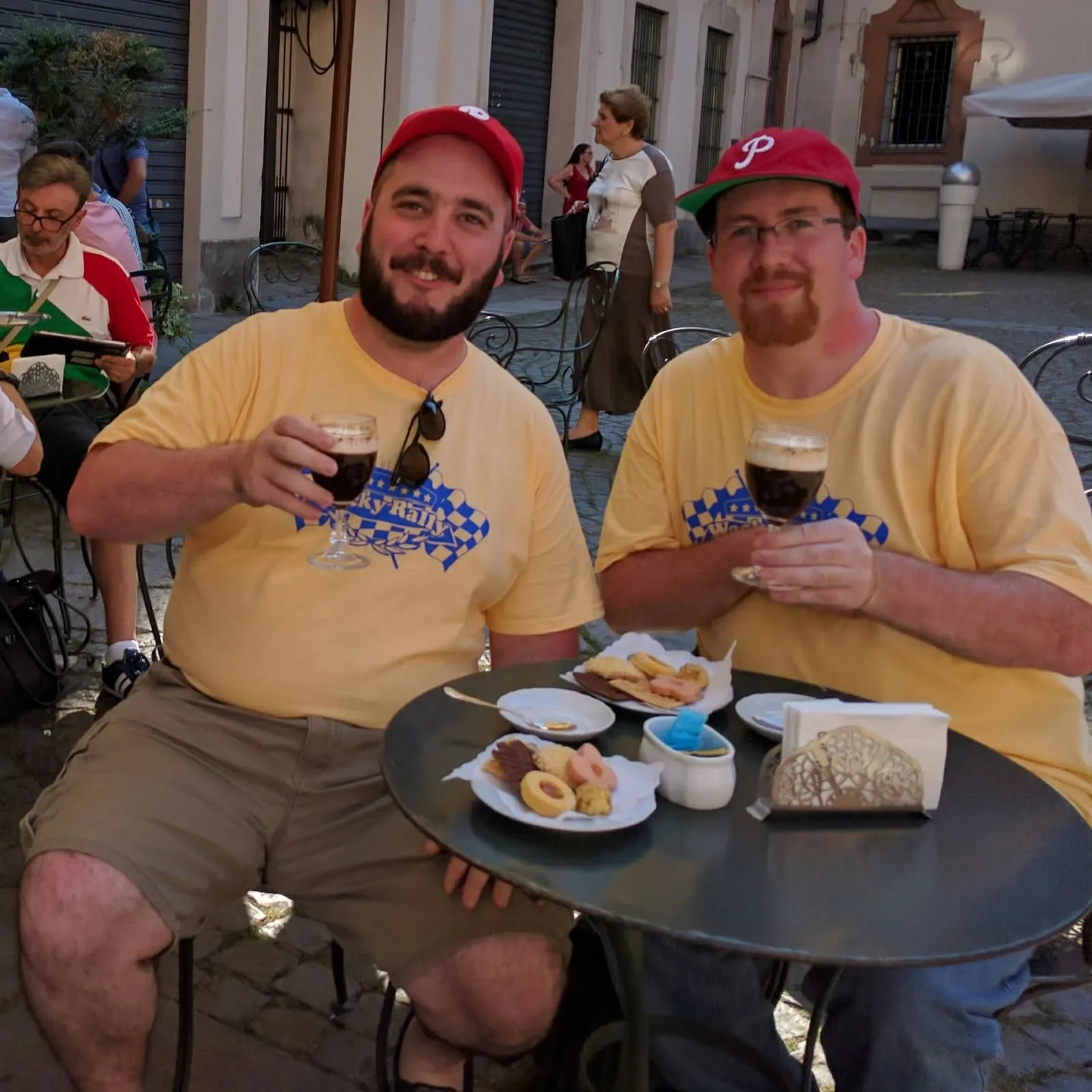 Two men wearing yellow shirts and red baseball caps sitting at an outdoor table, holding glasses of dark beer, with plates of snacks and condiments in front of them, at a café or restaurant.