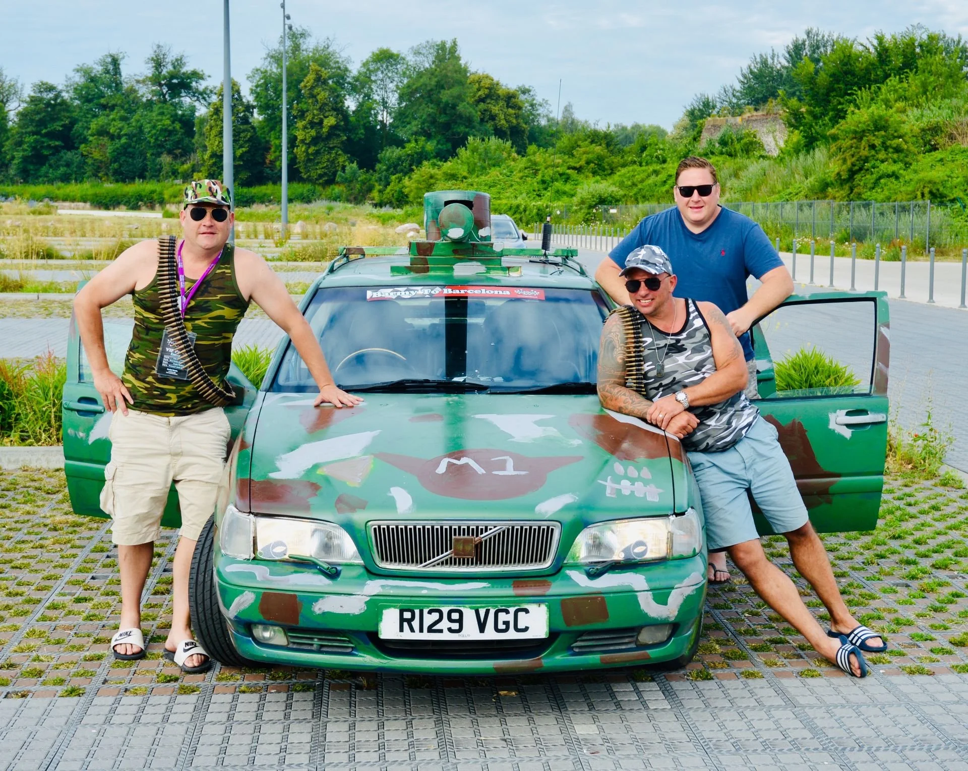 Three men with sunglasses posing with a camouflage-painted car outdoors on a paved area, with trees and a fence in the background.