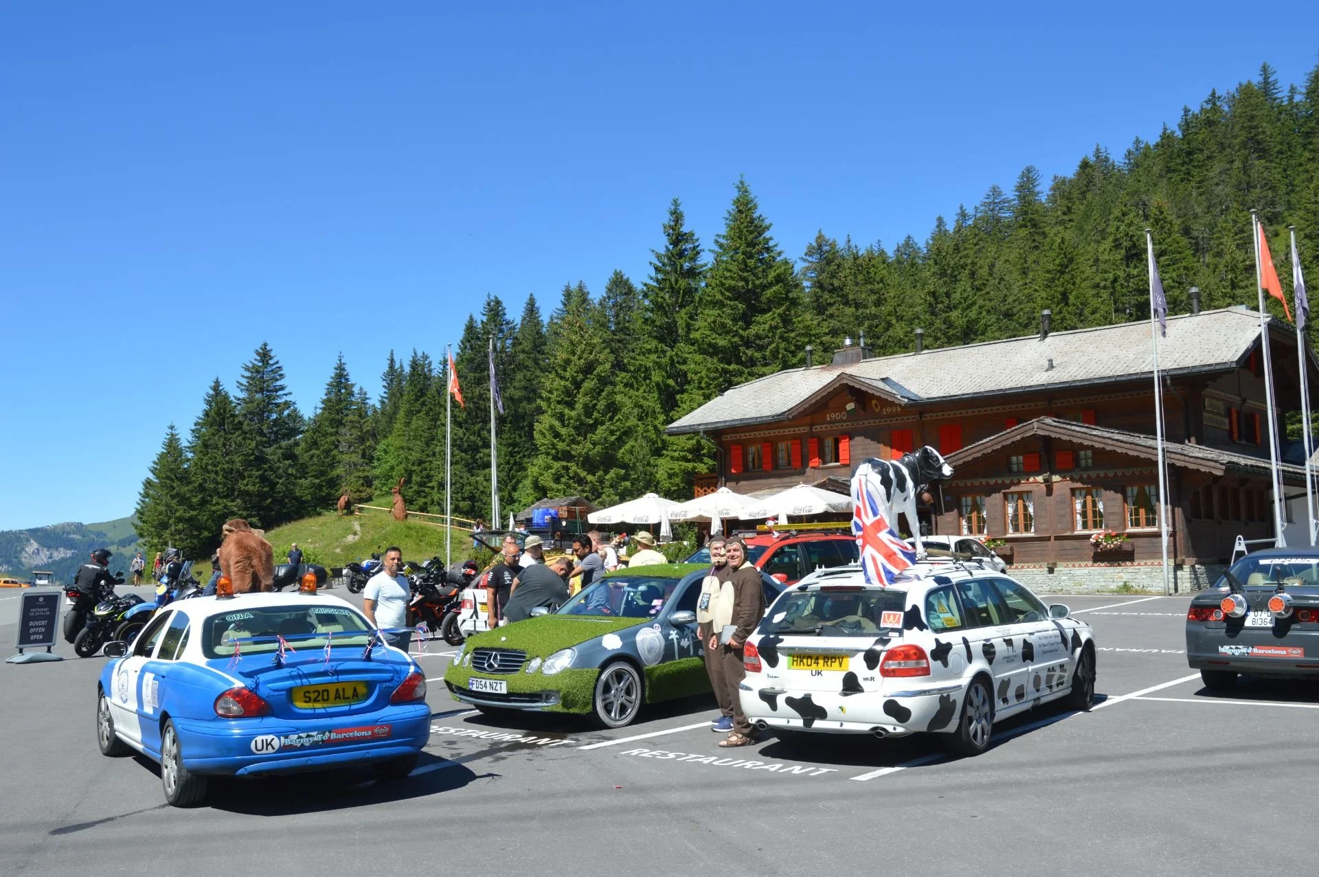A parking lot filled with decorated cars and people outside a chalet-style building amid a lush green forest under a blue sky.