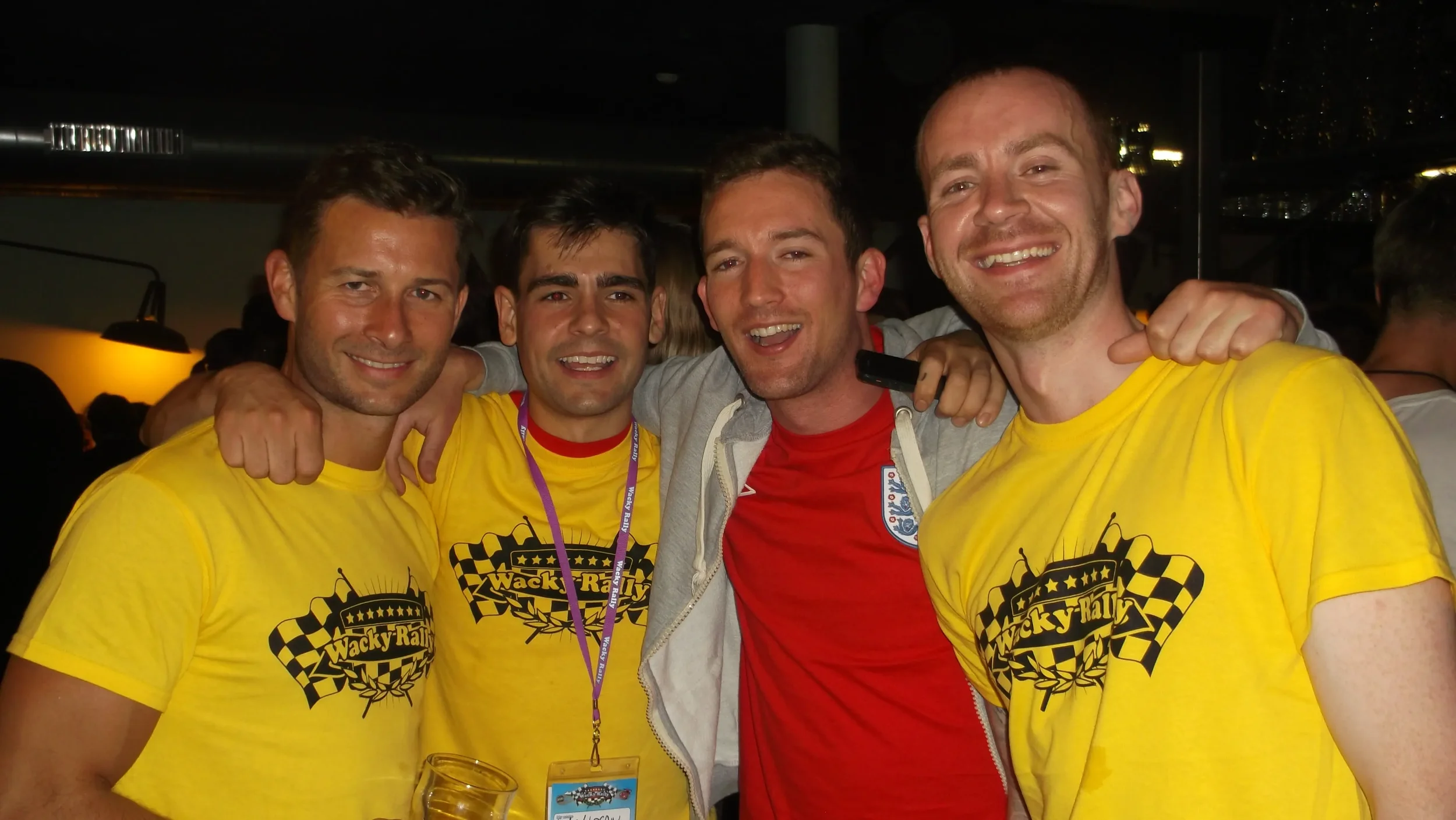 Four men smiling and posing together at an indoor event. Two of them are wearing yellow T-shirts with a race-themed logo that reads 'Wacky Races', and one is wearing a red shirt with England's national team emblem. They have their arms around each ot