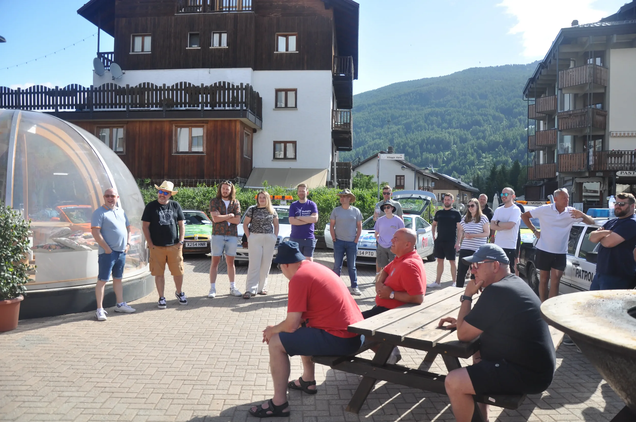 Group of people gathered outdoors in a small town square, standing and sitting around during daytime, with multi-story buildings, parked cars, and mountainous landscape in the background.