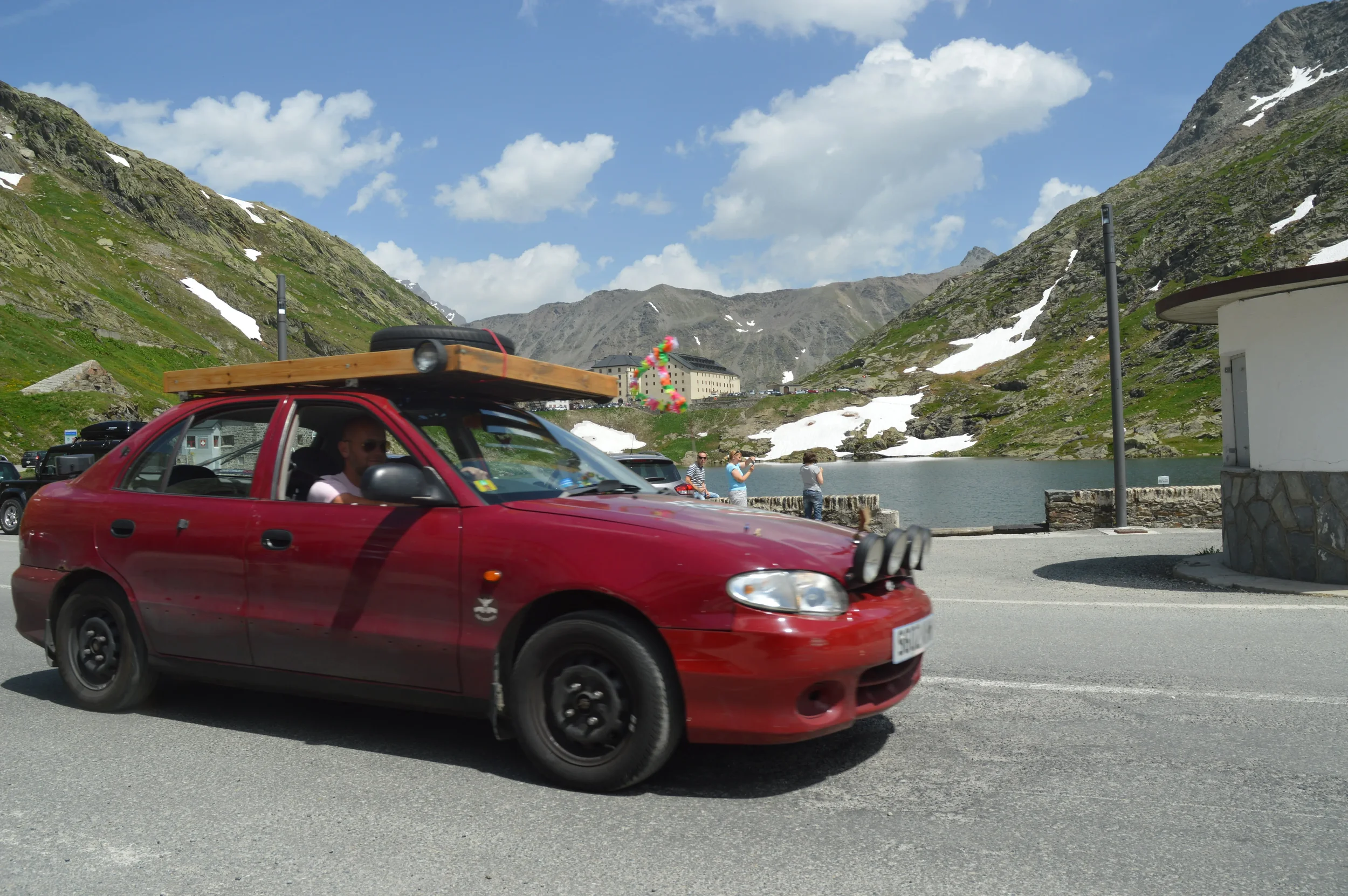 Red car with wooden roof rack and equipment, driving near a mountain lake with snow patches, with people taking photos in the background.