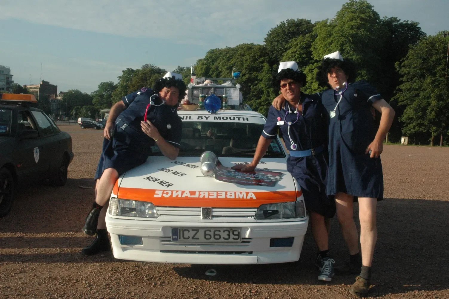 Three people dressed in nurse costumes with wigs and stethoscopes posing in front of a makeshift ambulance vehicle, with a parking lot and trees in the background.