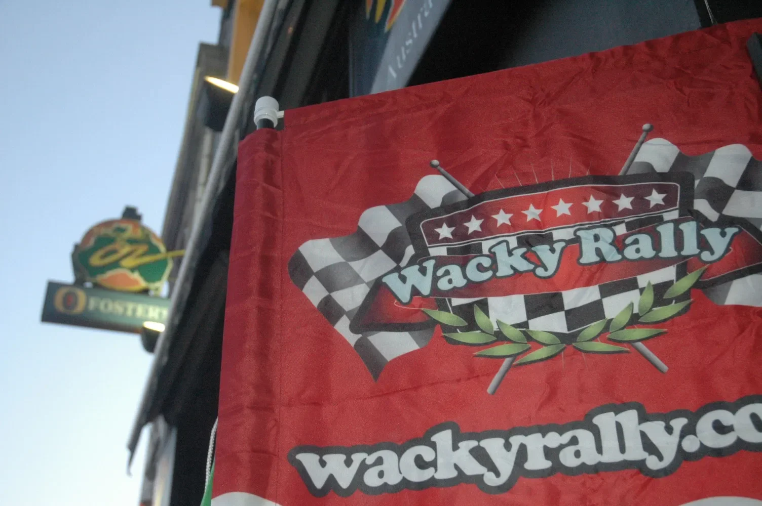 Red banner with 'WackyRally' logo, checkered race flag pattern, and green laurel leaves, hanging in front of a building with a sign for a motel or restaurant in the background.