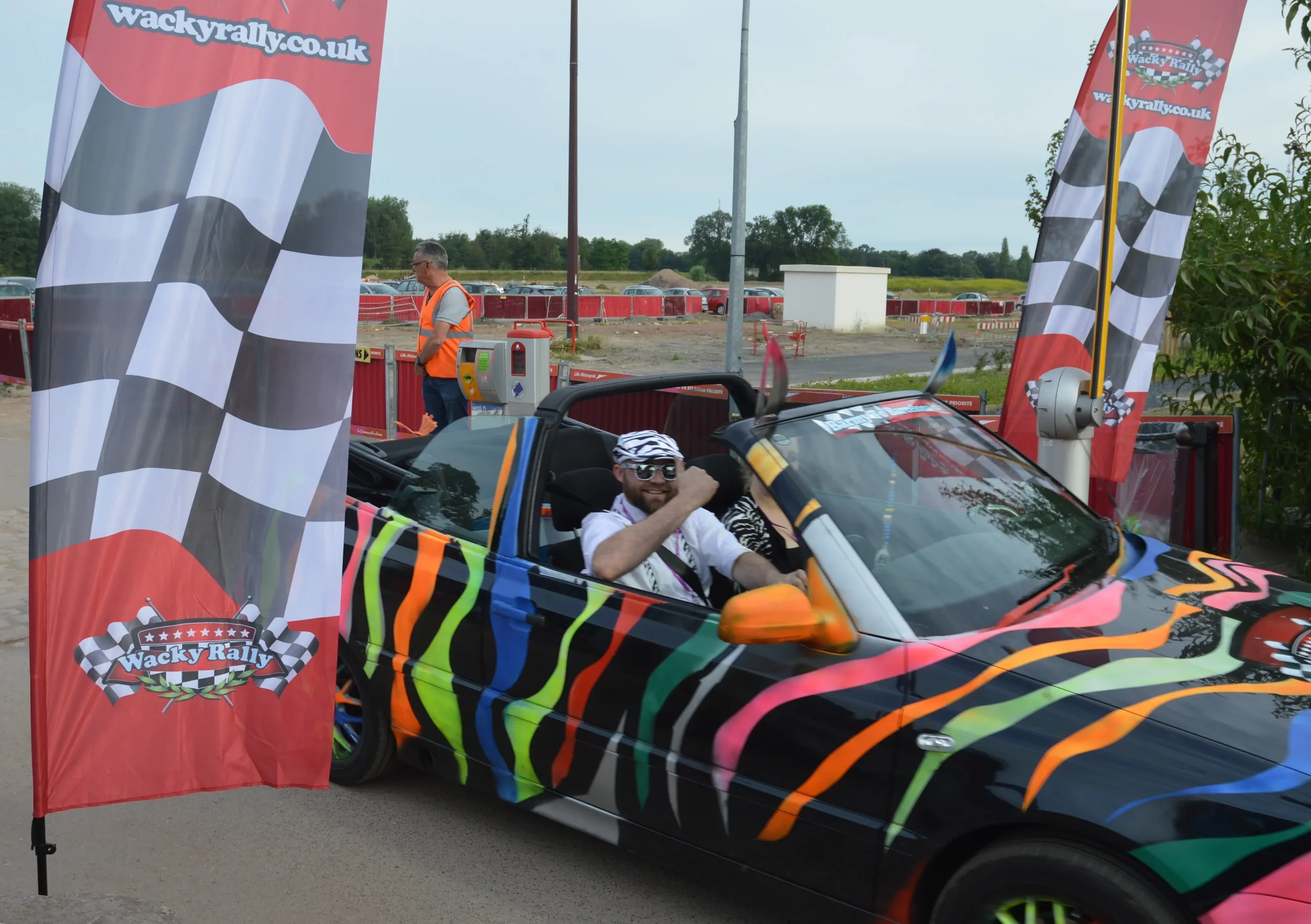 A man with sunglasses, a white shirt, and a zebra stripe bandana in a black sports car with colorful rainbow stripes, giving a thumbs up. The car is at a finish line with red flags that say 'Wacky Rally' and checkered pattern, and a man in an orange 