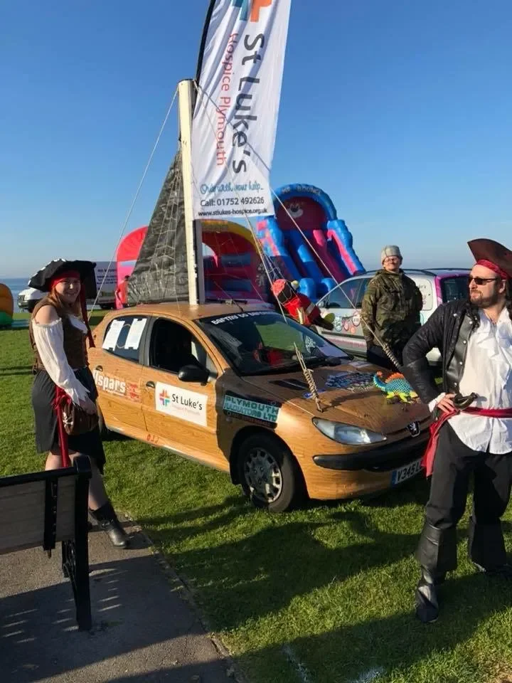 A display at an outdoor event featuring a car decorated to resemble a pirate ship, with a large sail, a slide, and people dressed in pirate costumes.