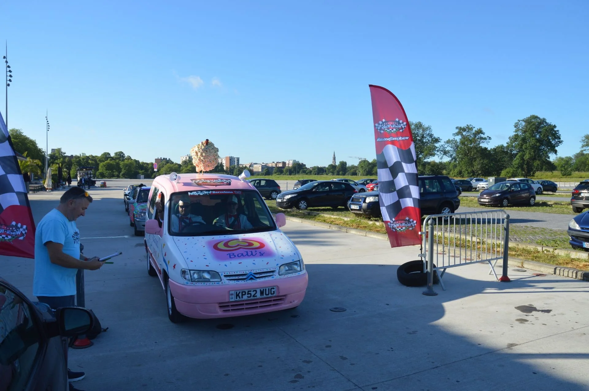 Pink car with a large popcorn decoration on top, parked outdoors during the day with flags, barriers, and other vehicles around. People are walking and looking at the car, indicating a car event or festival.