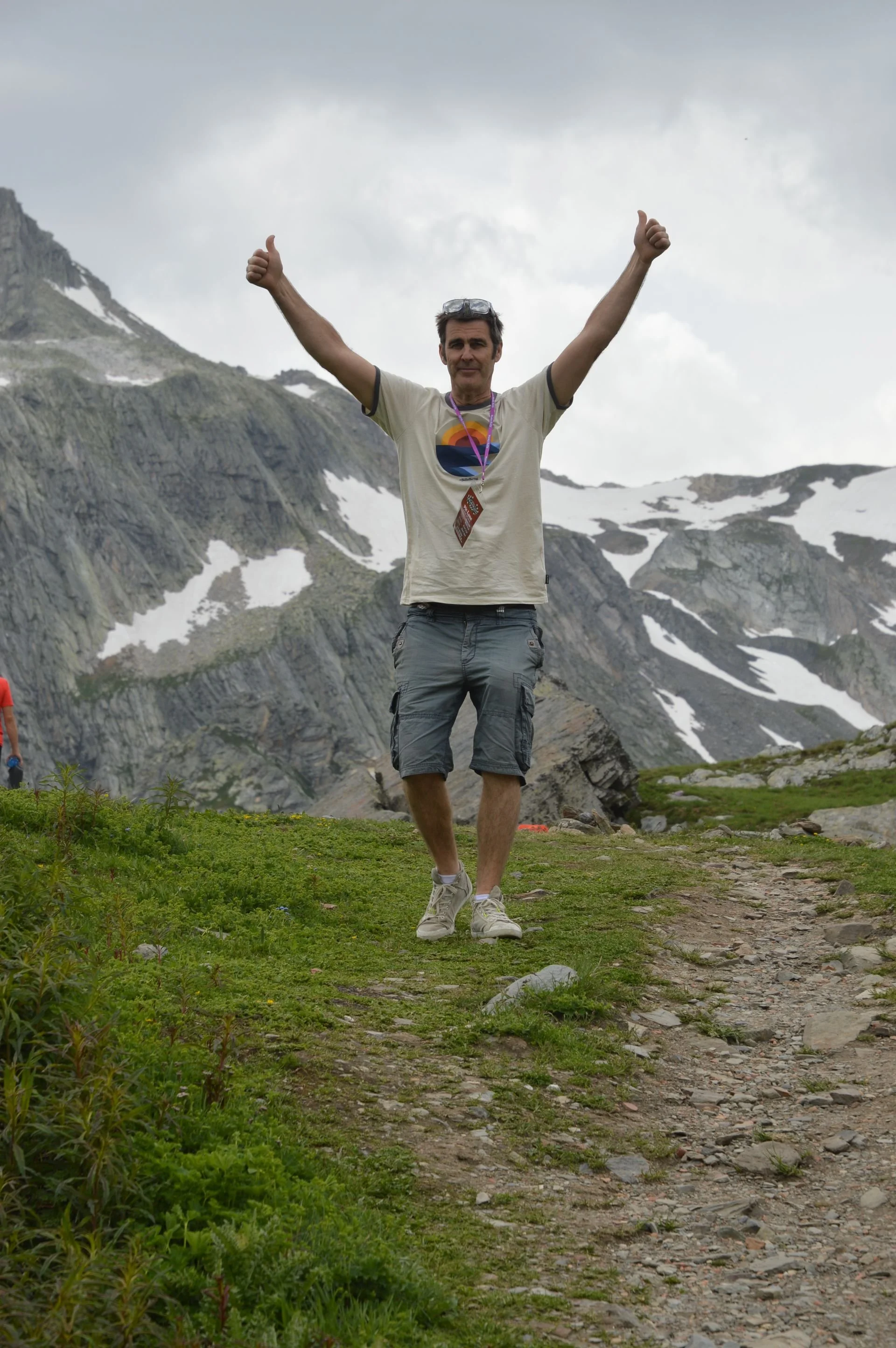 A man doing a dance move on a mountain trail with mountains and snow patches in the background.