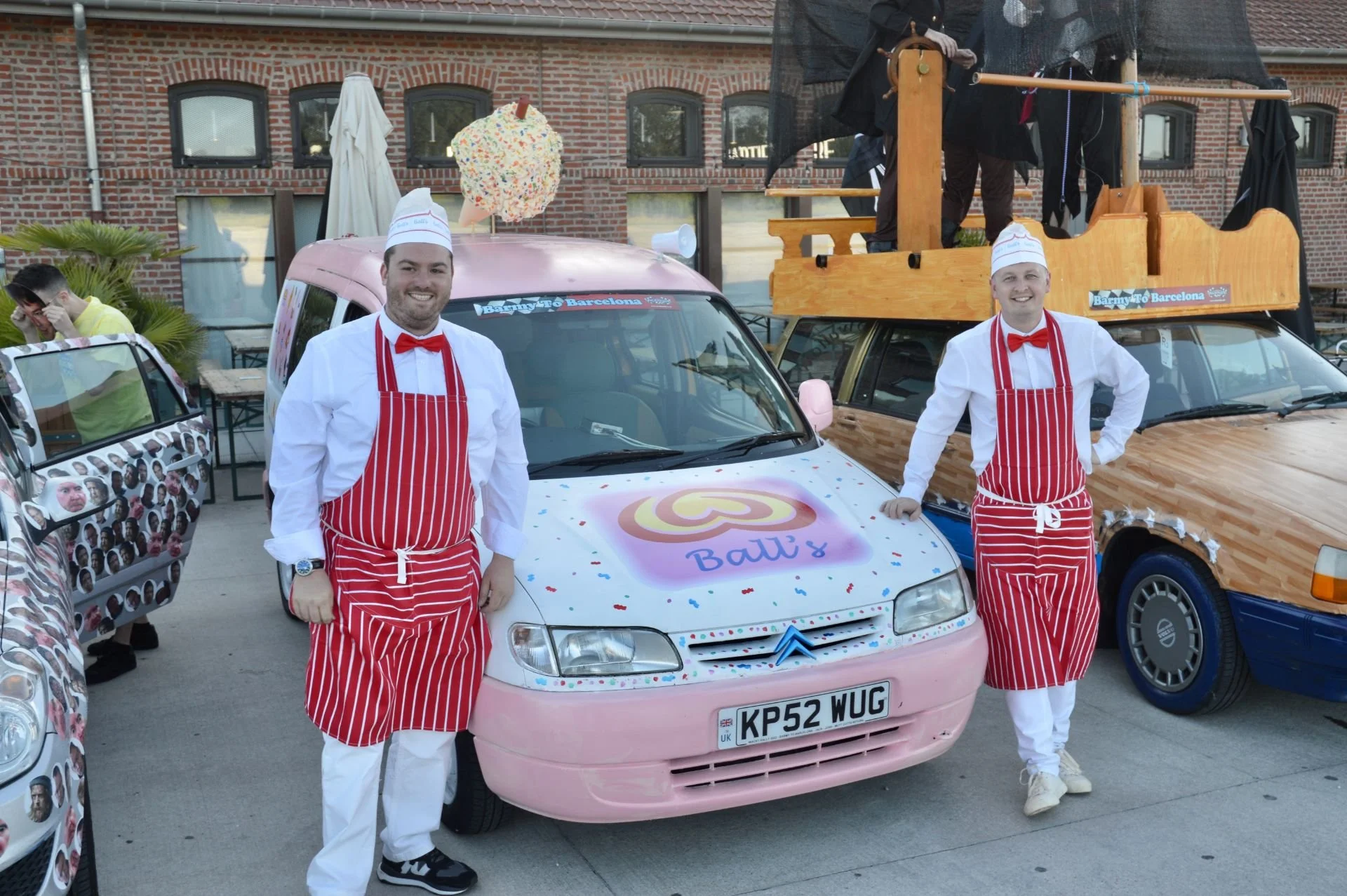 Two men dressed as ice cream vendors in red and white striped aprons and hats, standing in front of a decorated pink car with ice cream branding, in a busy outdoor setting with other customized cars and people.