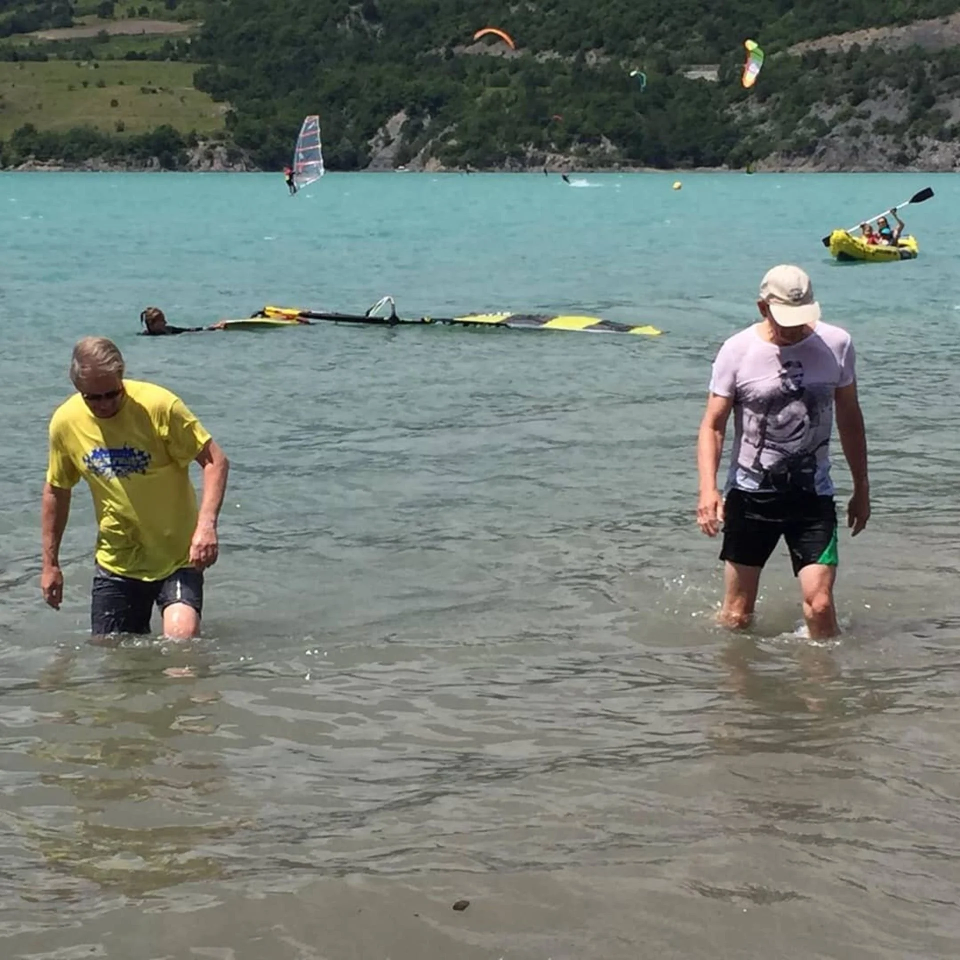 Two people standing in shallow water near the beach, with a partially submerged yellow boat in the background and a boat upside down in the water. Several kite surfers and a kayak are visible further out on the lake, with green hills and trees in the