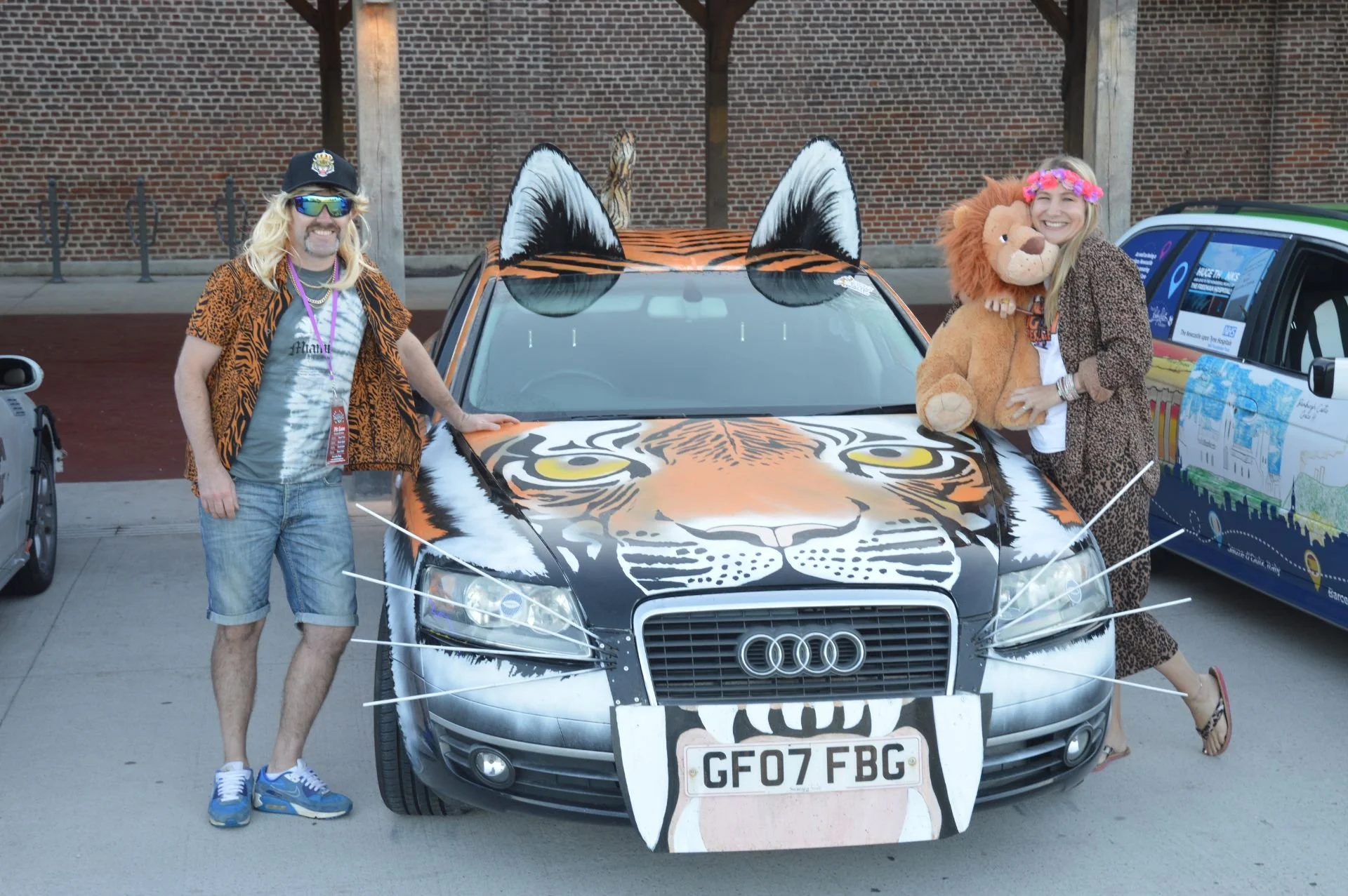 People dressed in animal-themed costumes posing next to a car decorated like a tiger, with a tiger face and ears on the hood and a tiger tail on the roof, in what appears to be a parking lot.