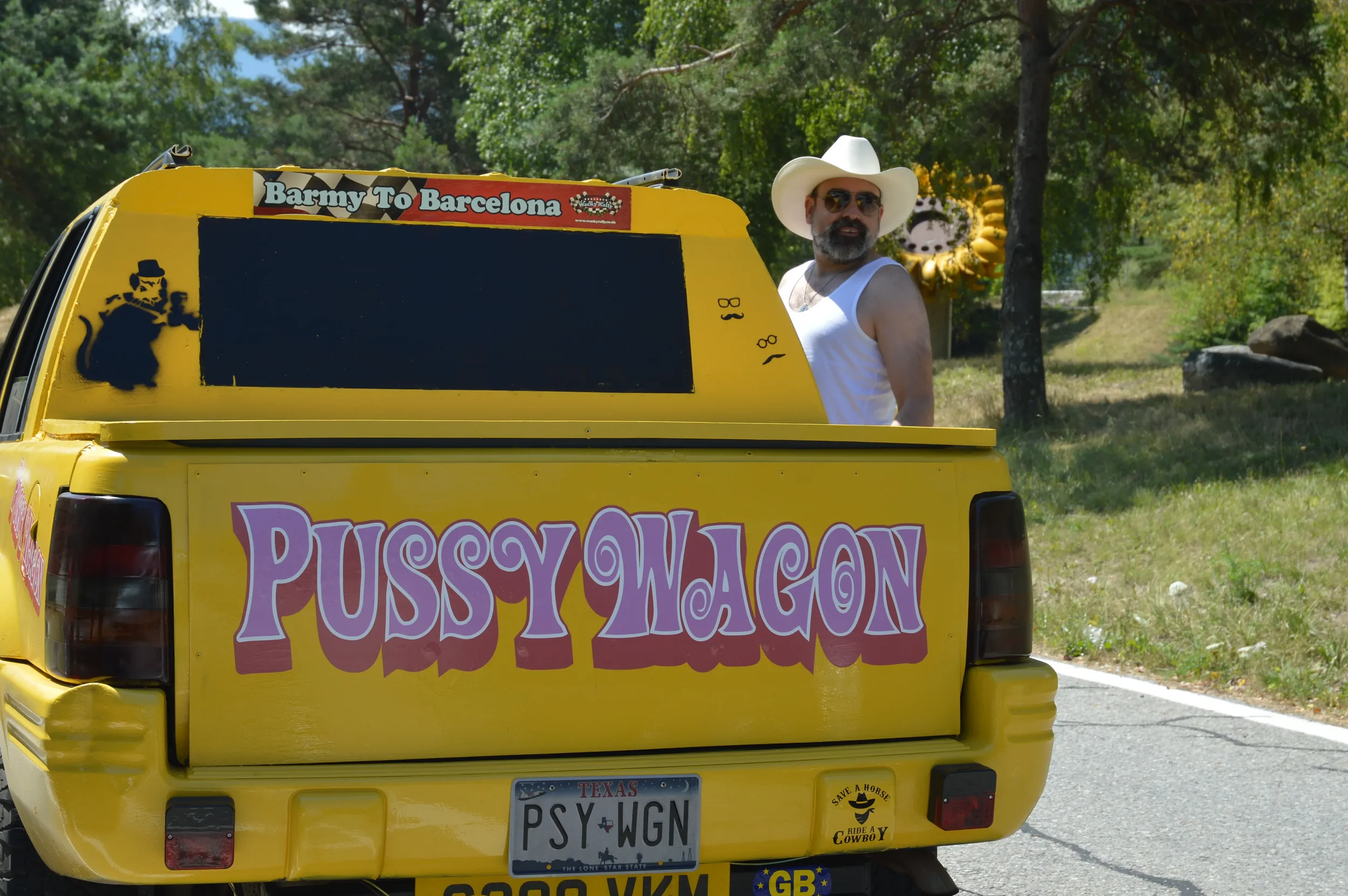 A man wearing a white cowboy hat, sunglasses, and a white sleeveless shirt stands next to a yellow vehicle labeled "Pussy Wagon" with a Texas license plate "PSY WGN". The vehicle has various stickers, including one that says "Barmy To Barcelona" and 