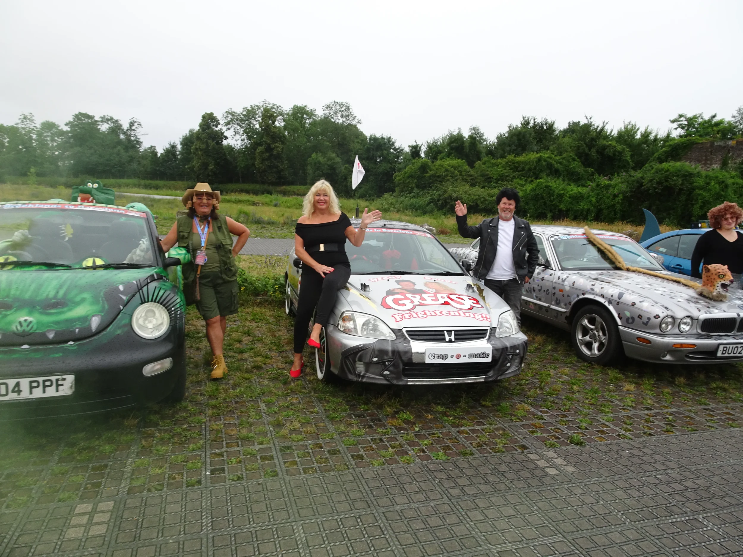 Four people posing with creatively decorated cars outdoors on a cloudy day, with trees in the background.