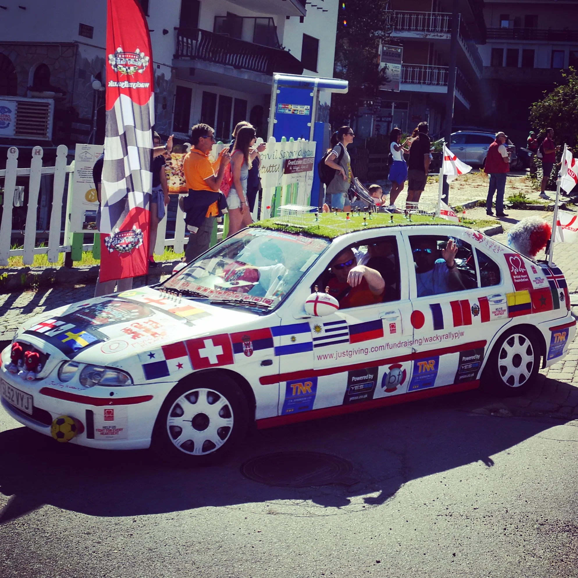 Decorated rally car with flags and sponsors, parked on street during event, with people standing in line and observing in the background.