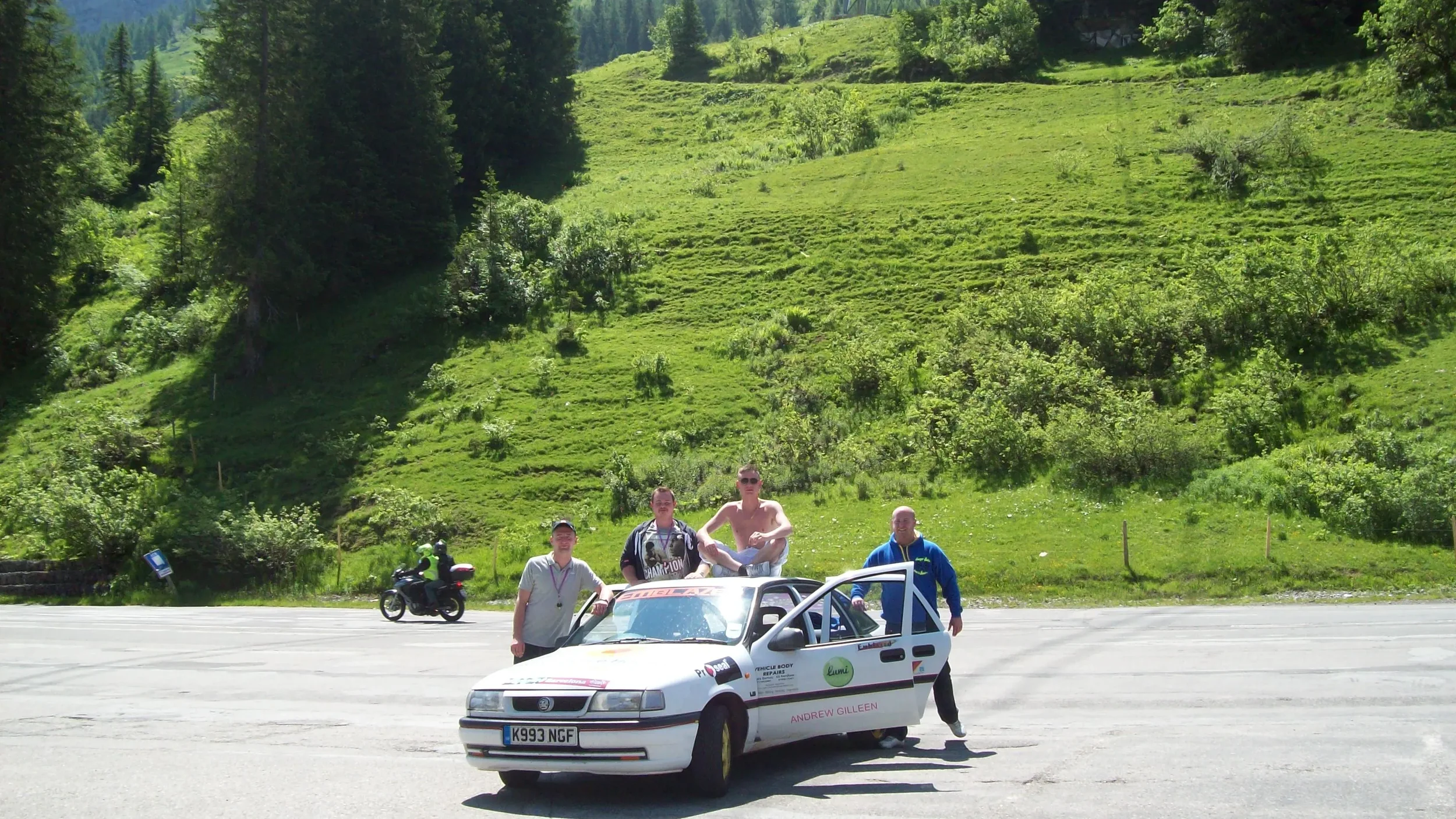 Four men posing with a white rally car on a mountain road, with a lush green hillside and trees in the background. One man is sitting on the roof, and the others stand near the car.