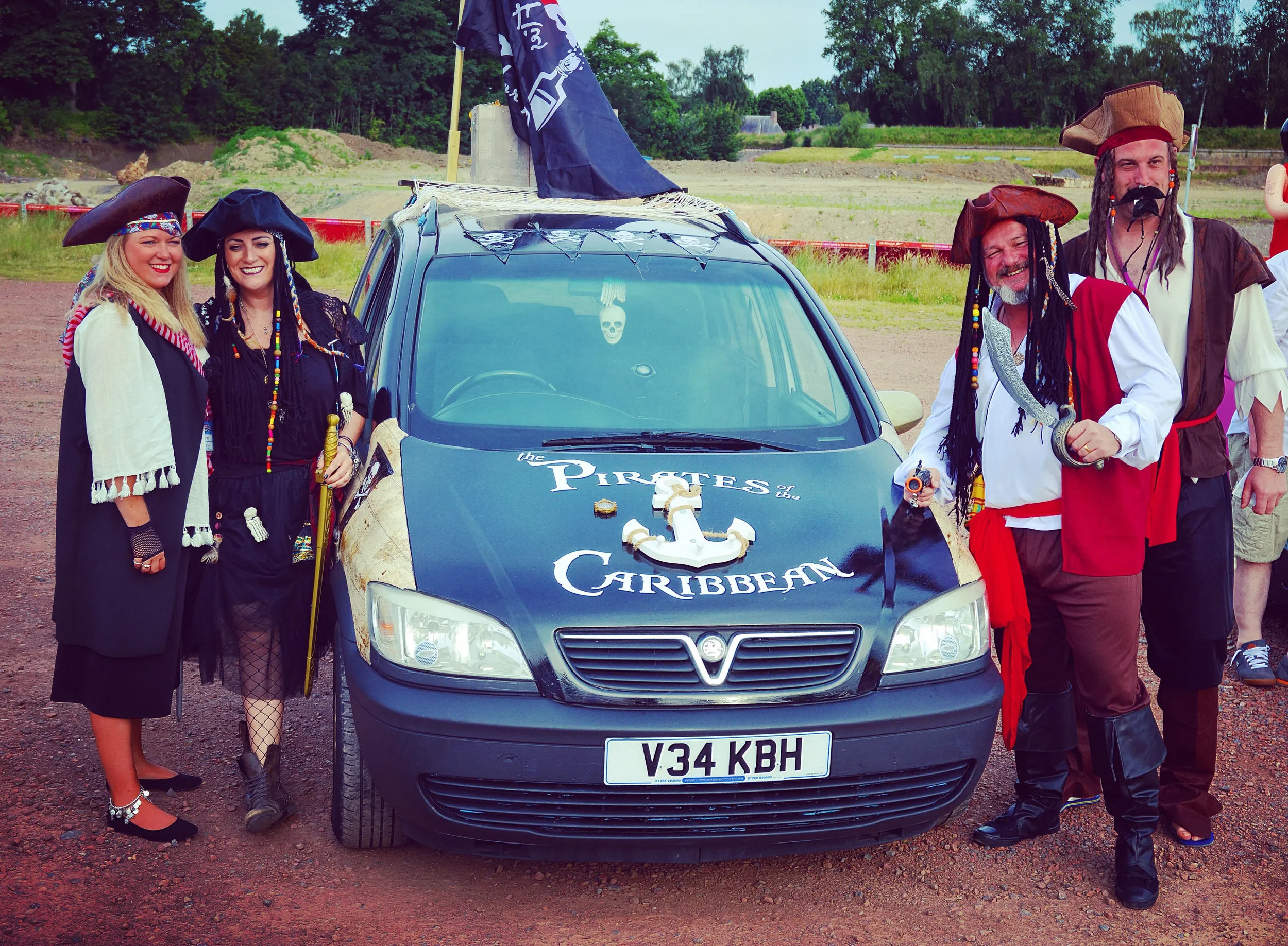 Group of five people dressed as pirates standing next to a decorated car with a pirate theme, including a skull and crossbones flag and the words 'Pirates of the Caribbean' on the hood.