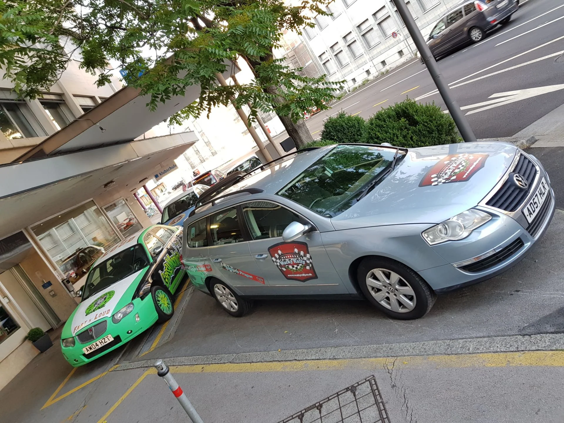 Two cars parked on the street, one green and one silver, with racing themed decals, situated near a building with large windows and a sidewalk. Trees and a street with moving vehicles are visible in the background.
