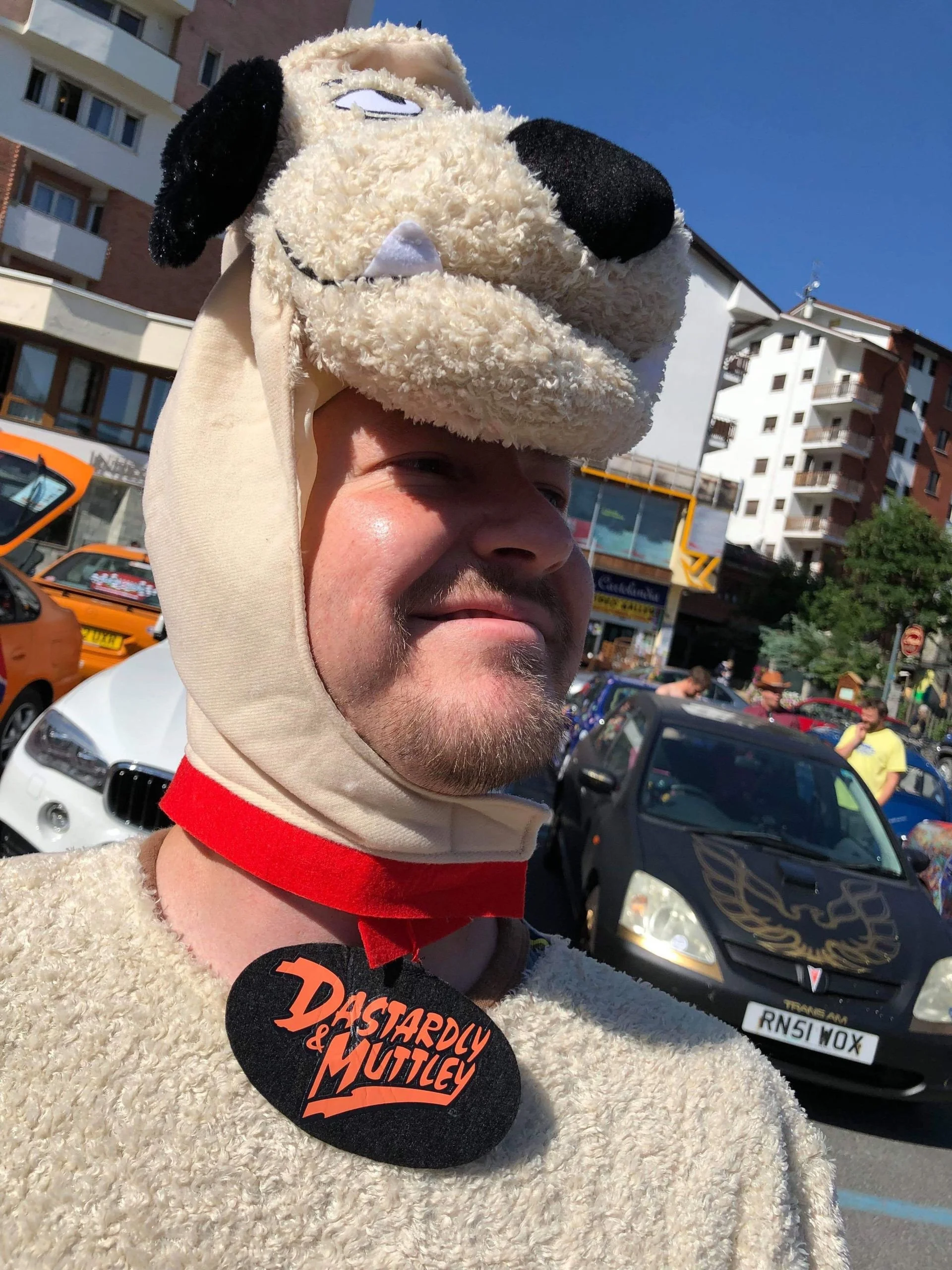 A person wearing a cartoon dog costume headpiece and a costume with a 'Dastardly & Muttley' badge, standing outdoors on a sunny day with buildings, cars, and people in the background.