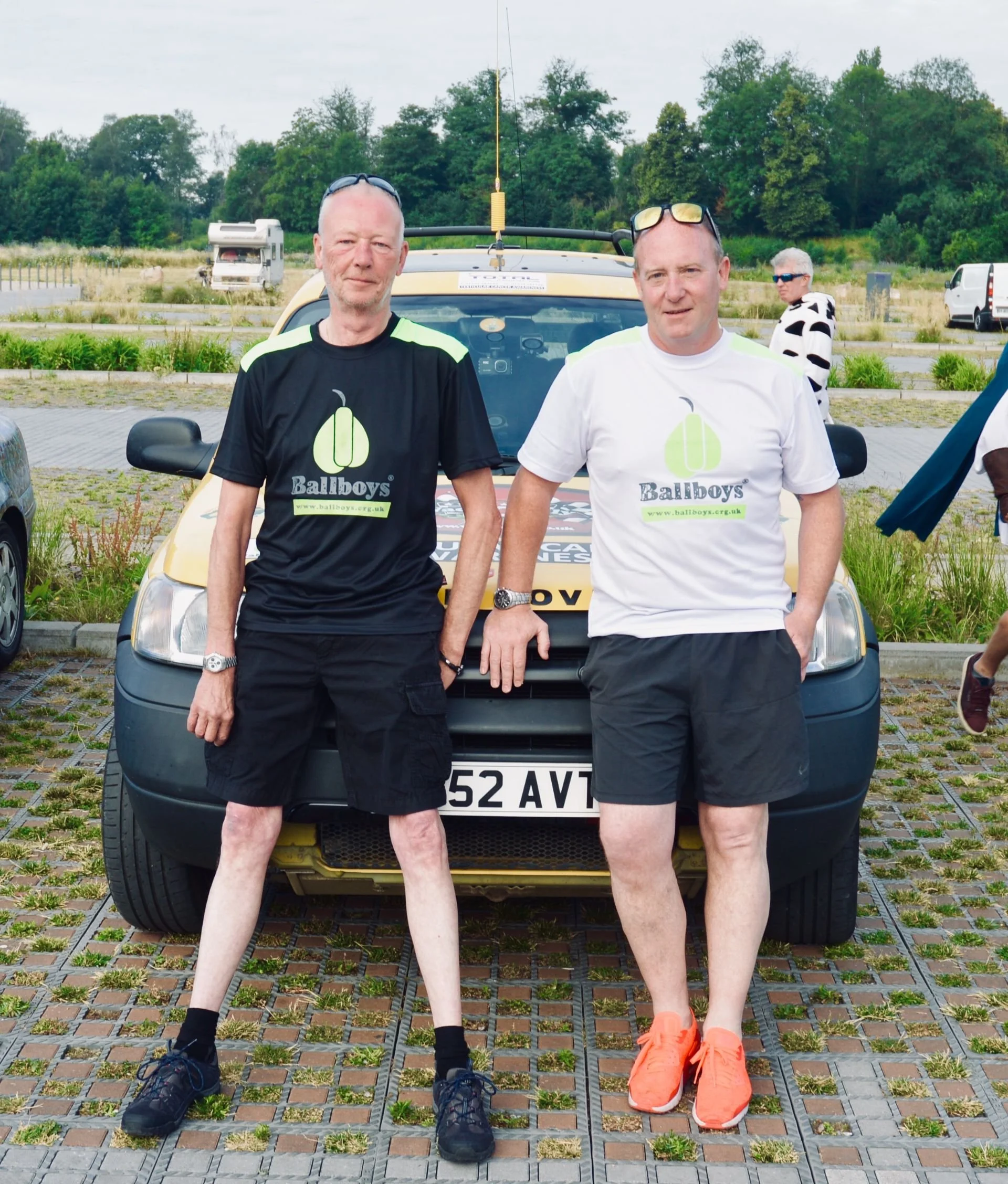 Two men standing in front of a yellow car, both wearing black and white t-shirts with a green logo and the word 'Ballboys,' in an outdoor parking lot with trees and other people in the background.