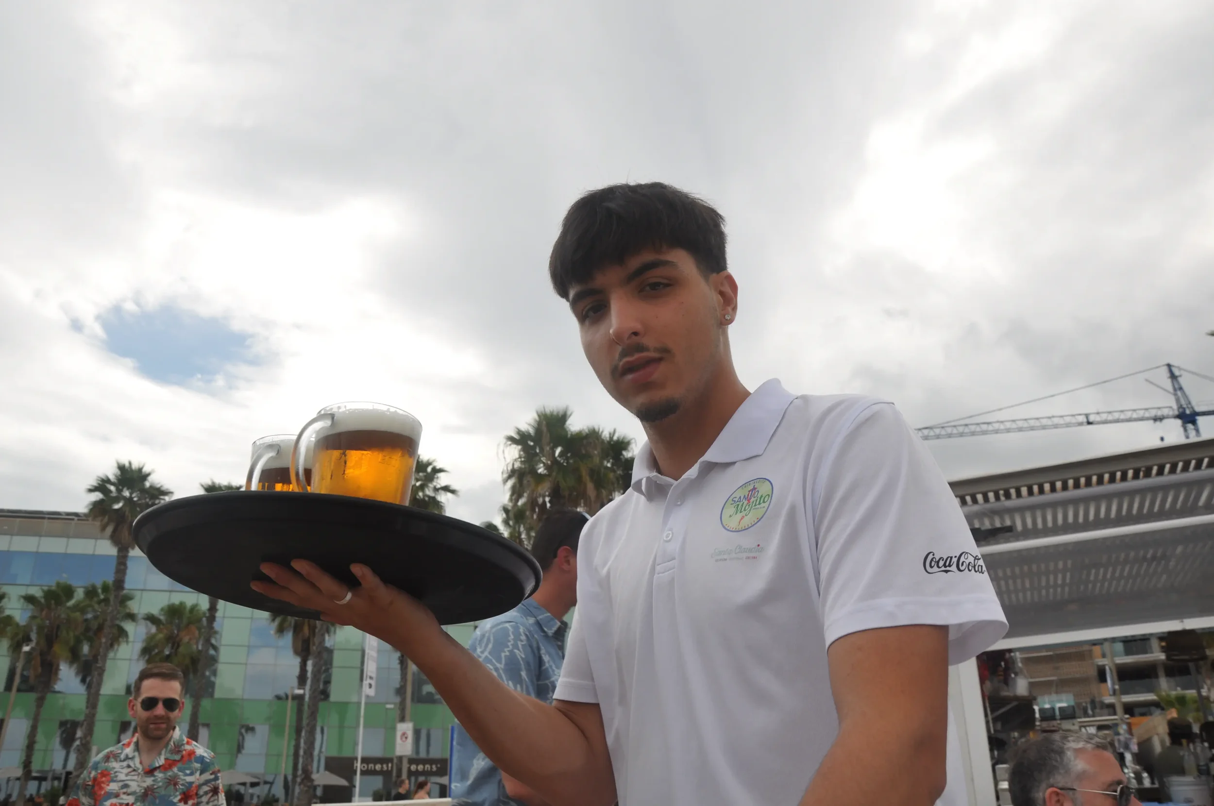 A young man in a white shirt holding a tray with two glass mugs of beer at an outdoor event, with palm trees and a modern building in the background.
