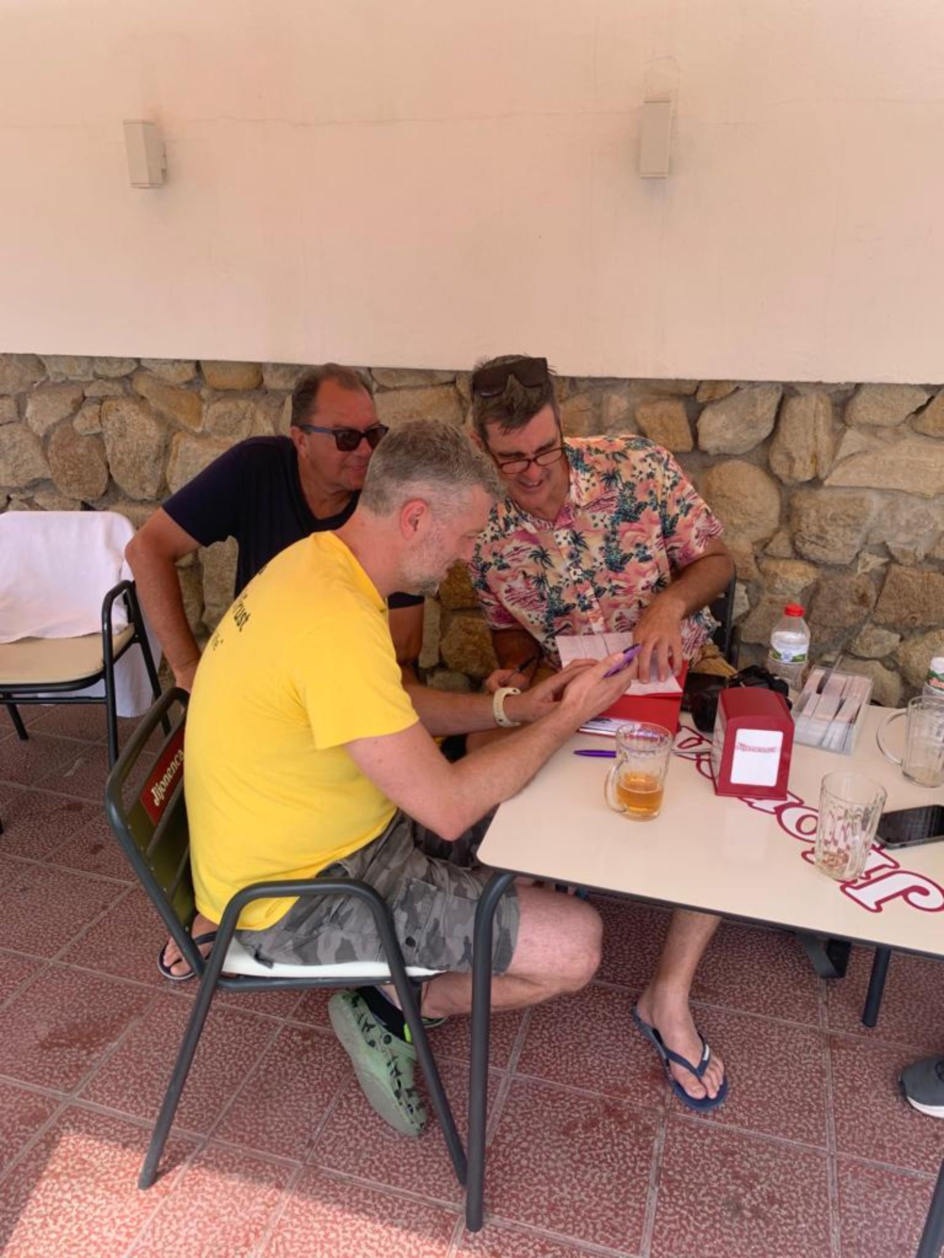 Three men sitting and standing around a table looking at a phone indoors, with drinks and items on the table, stone wall background.