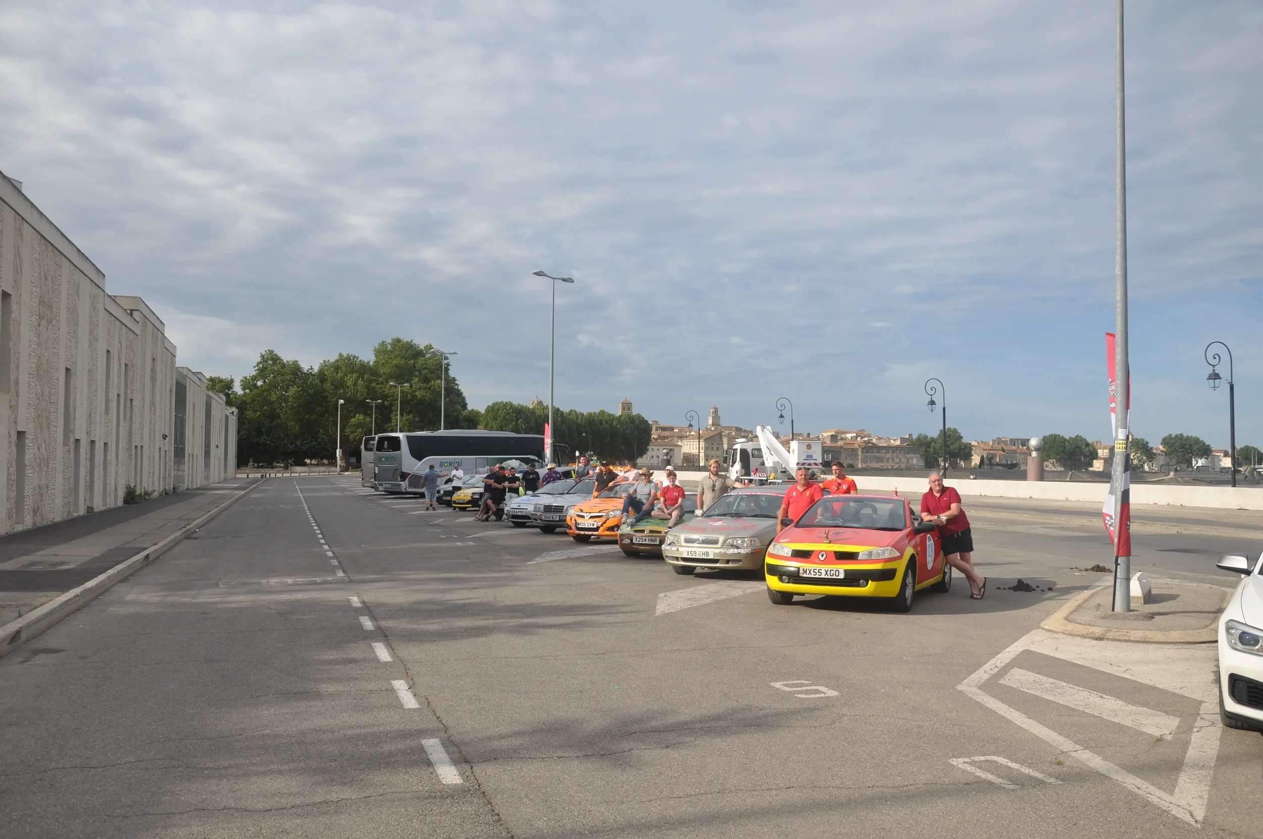 A lineup of cars and buses parked in a lot with people standing around, some leaning on cars, under a partly cloudy sky. There are street lamps and a white wall on the left, with a cityscape with historic buildings in the background.