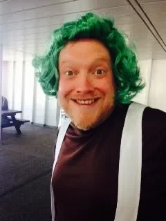 Person wearing a bright green curly wig, smiling, in an indoor setting with a backpack strap visible, and a bench in the background.