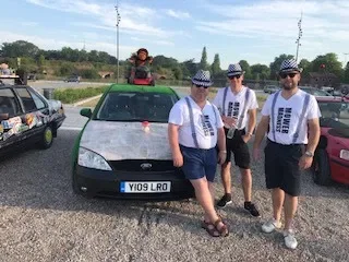 Three people standing in front of a silver car, wearing matching white t-shirts with barcode designs and sunglasses, in a parking lot.