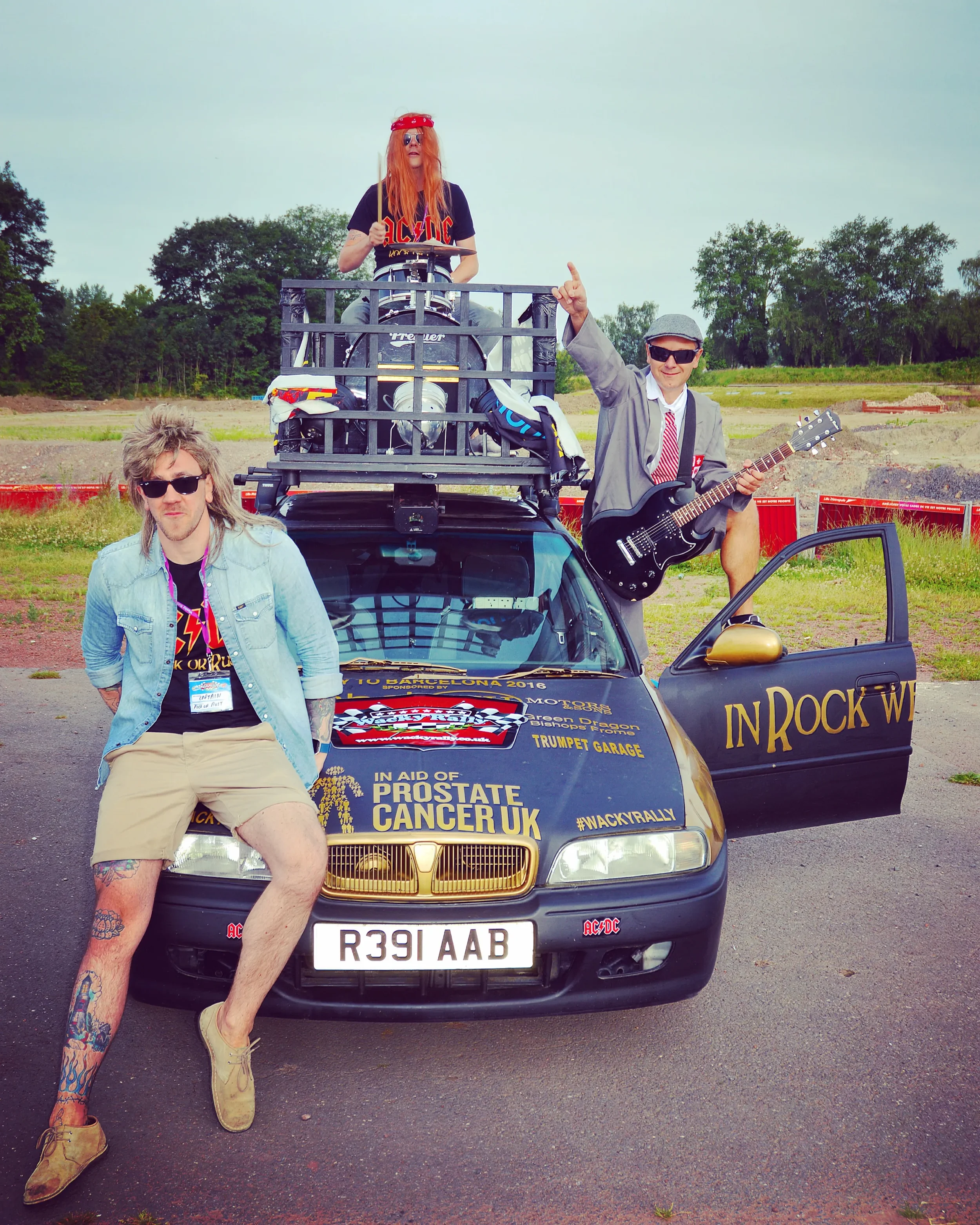 Three young men with tattoos and sunglasses posing around a black car with 'In Rock We Trust' written on the door, decorated for a charity event, with one on top of the car playing drums, another on the ground leaning against the hood, and the third 