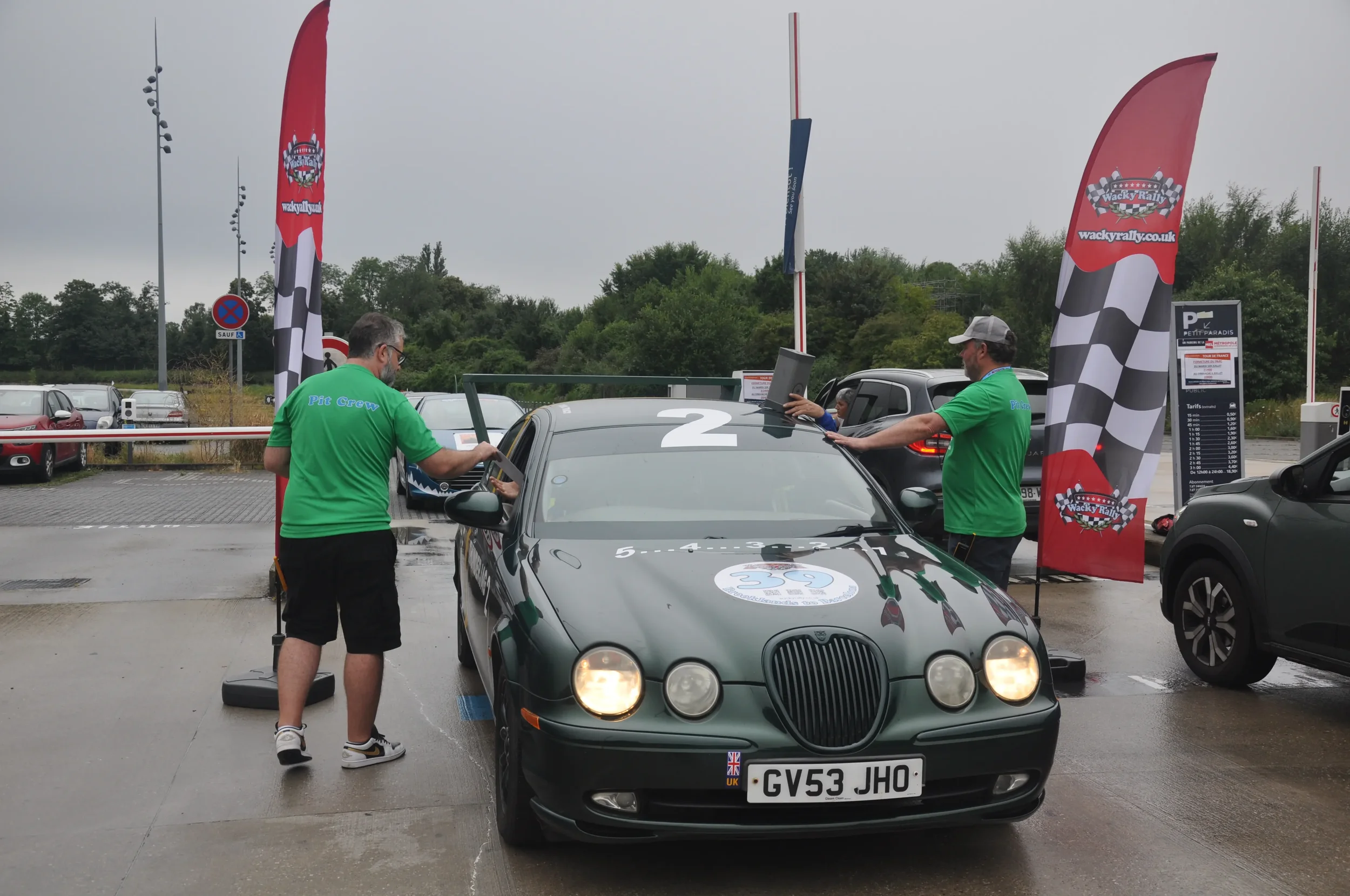 Car participating in a robot race event, with two men in green shirts using laptops and sensors at the front and the side of the car, which is parked in a parking lot with flags and signs around.