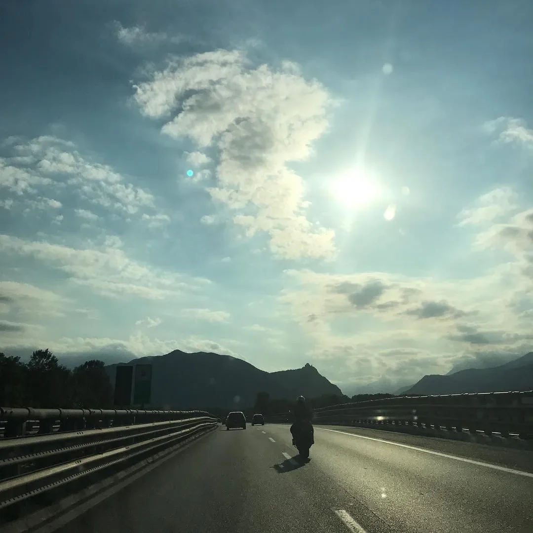 A highway with a motorcycle, a few cars, landscape mountains, and a partly cloudy sky with the sun shining through clouds.