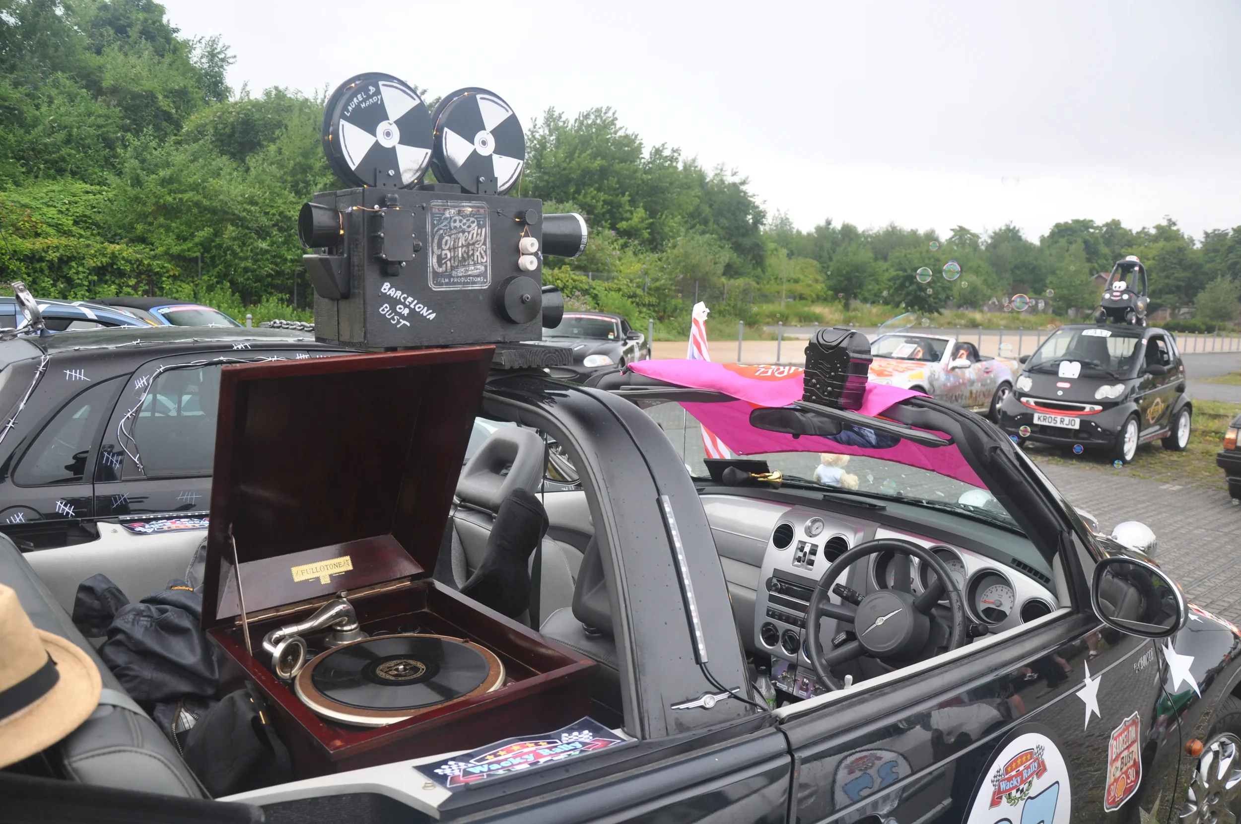 A black car with stickers and a vintage record player inside, equipped with a movie camera apparatus on top, surrounded by other cars at a gathering or event in a parking lot with green trees in the background.
