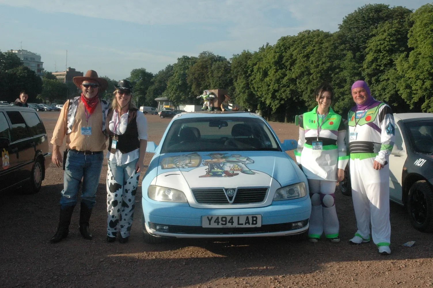 Group of five people dressed as characters from Toy Story standing in front of a light blue car with Toy Story characters painted on the hood at an outdoor event.