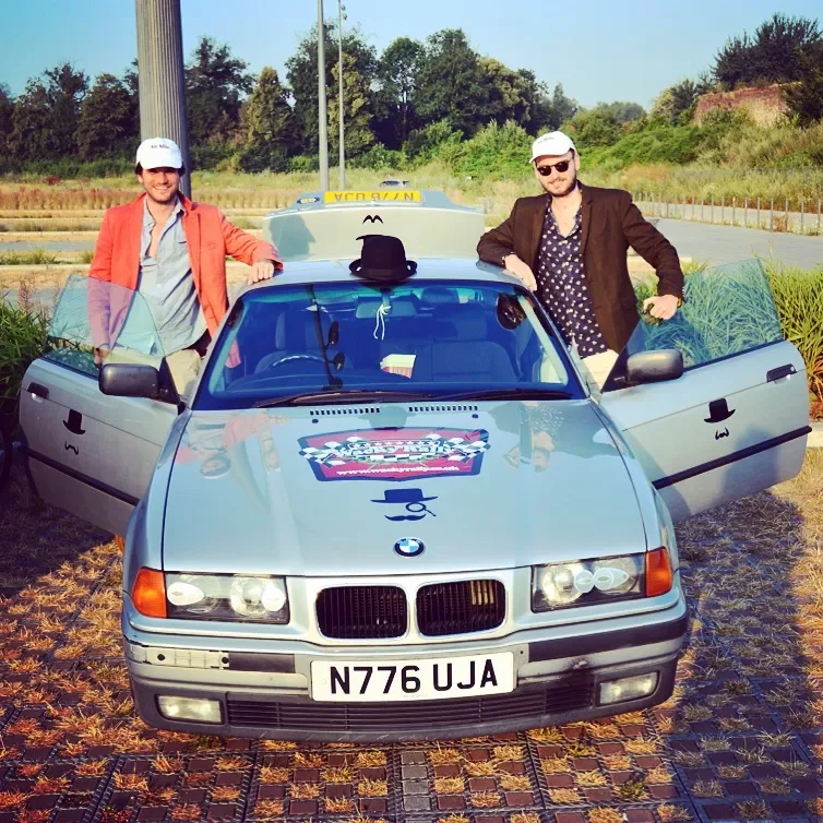 Two men standing beside a silver BMW car with taxi signs on the roof, open car doors, baseball caps, and sunglasses in an outdoor parking area.