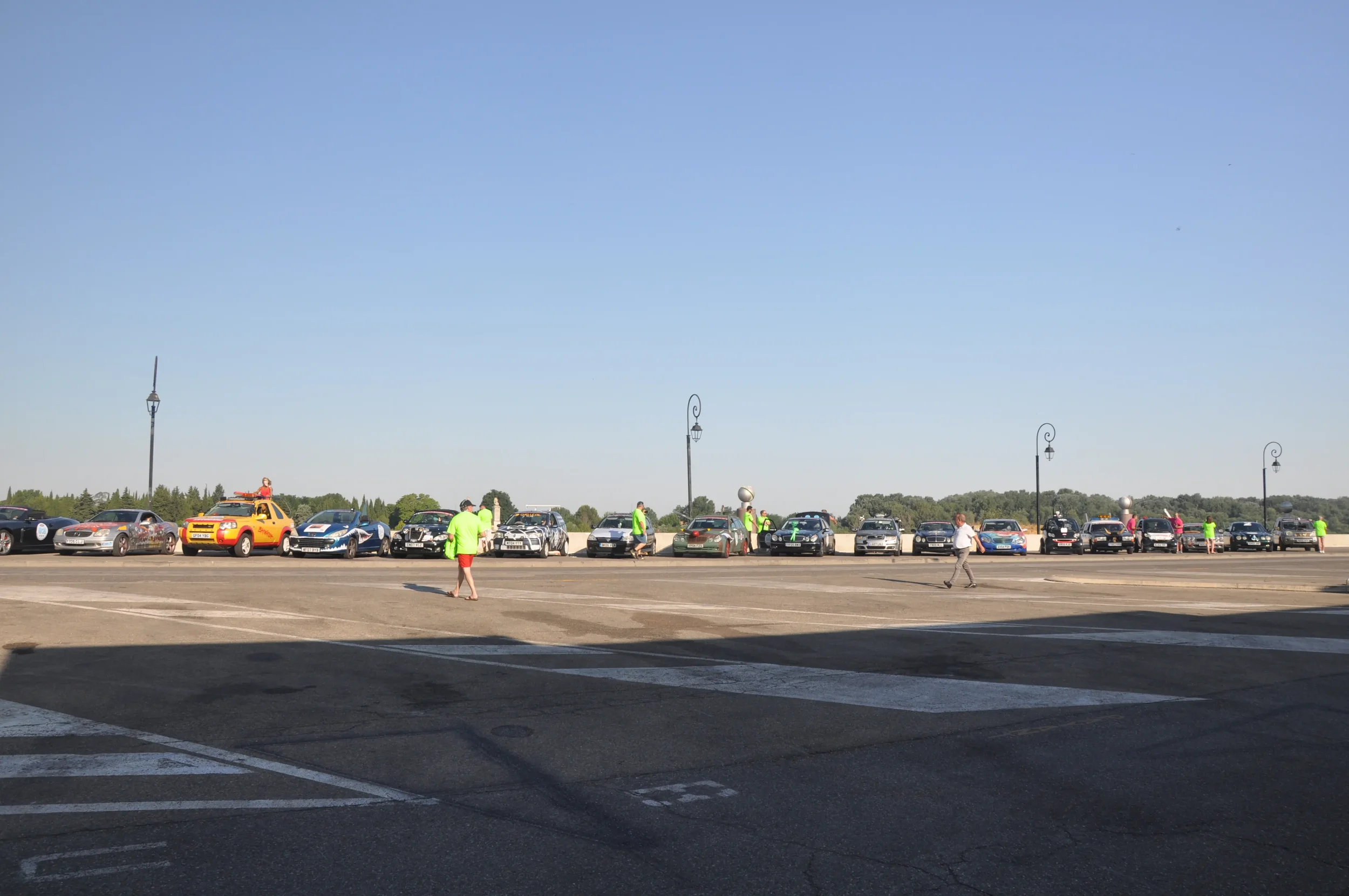 A row of parked cars on a large parking lot under a clear blue sky, with people walking and some in bright green shirts.