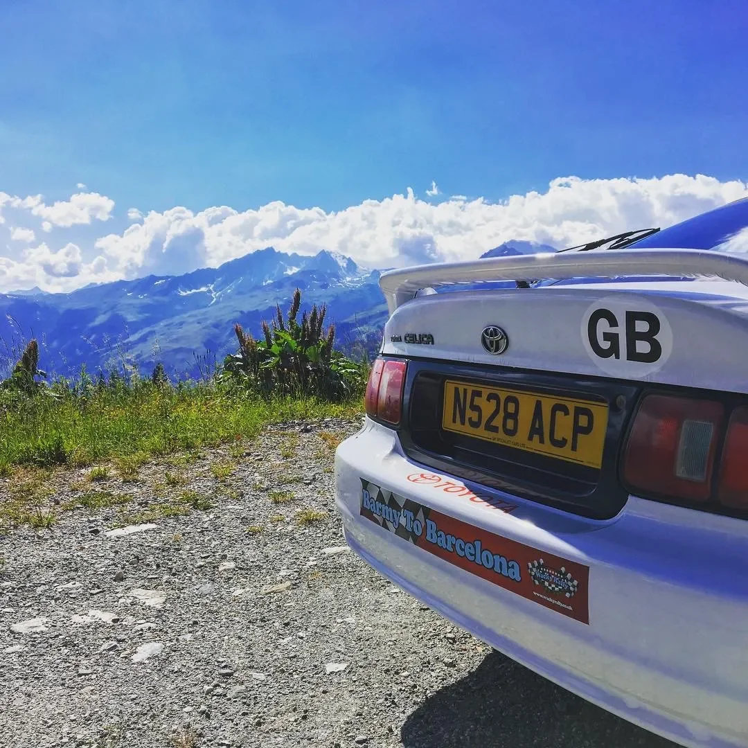 The back of a white Toyota Celica with a British license plate N528 ACP, parked on a gravel area with green grass and some tall plants, overlooking a mountain range under a partly cloudy blue sky. The car has a sticker that reads "Rally to Barcelona.