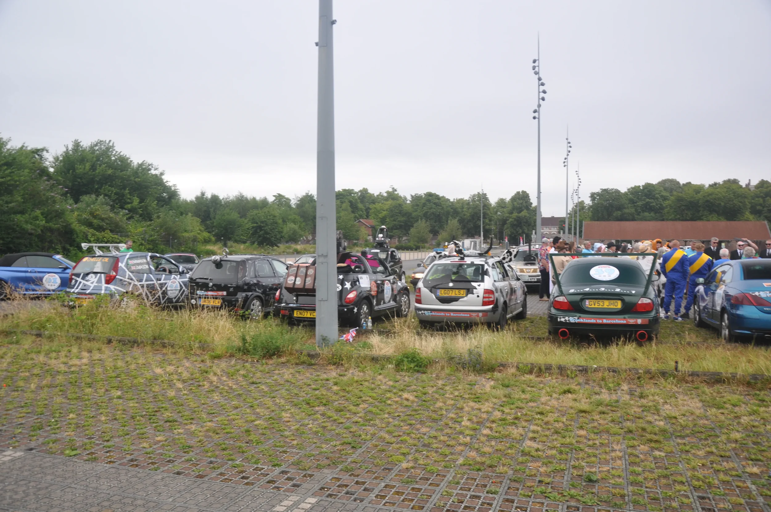 A parking lot filled with various decorated cars and a group of people gathered around, with some dressed in racing uniforms, under a cloudy sky.