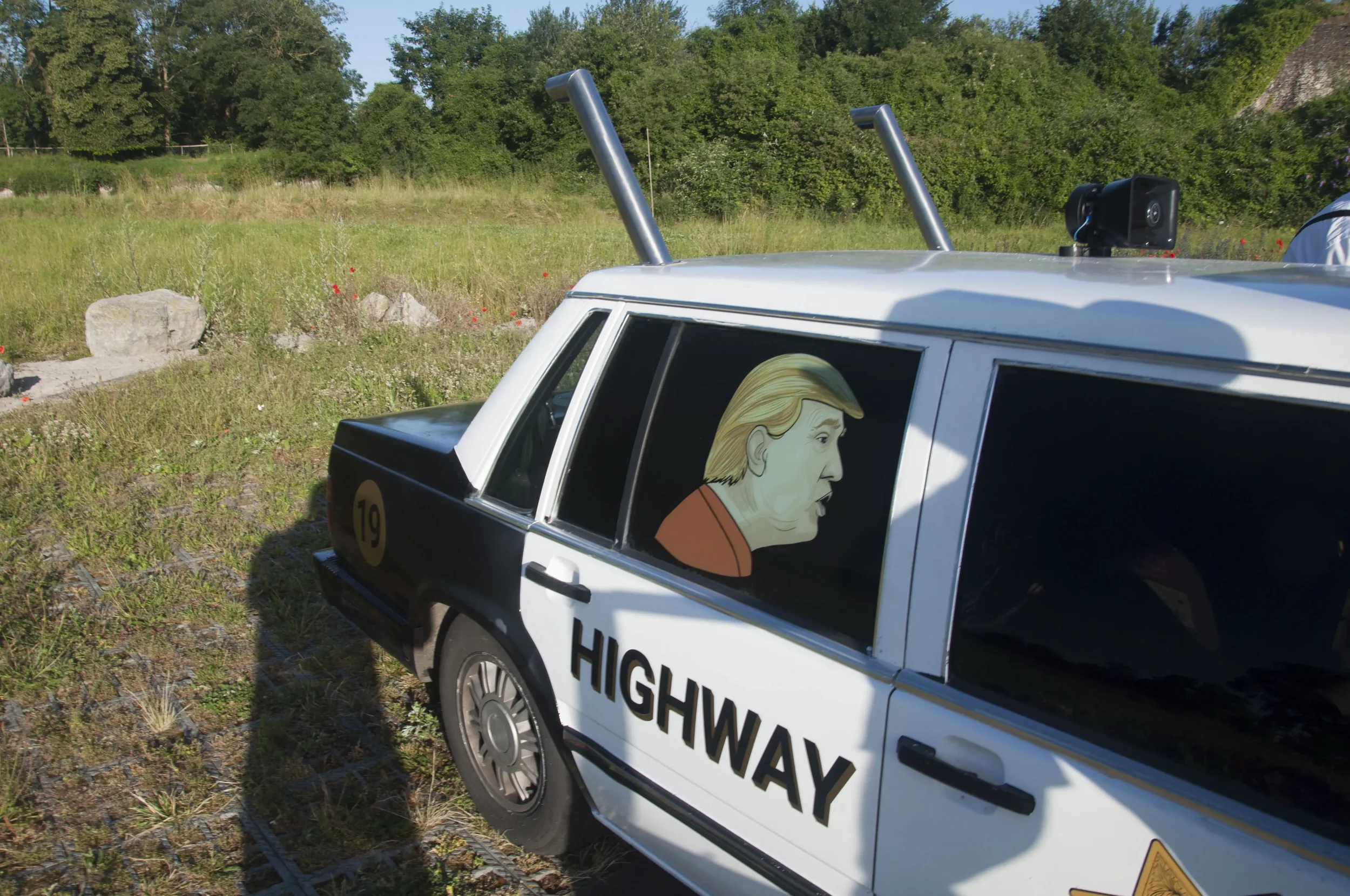 A police car with the fictional character caricature of Donald Trump on the side window, parked on grass with a field and trees in the background.