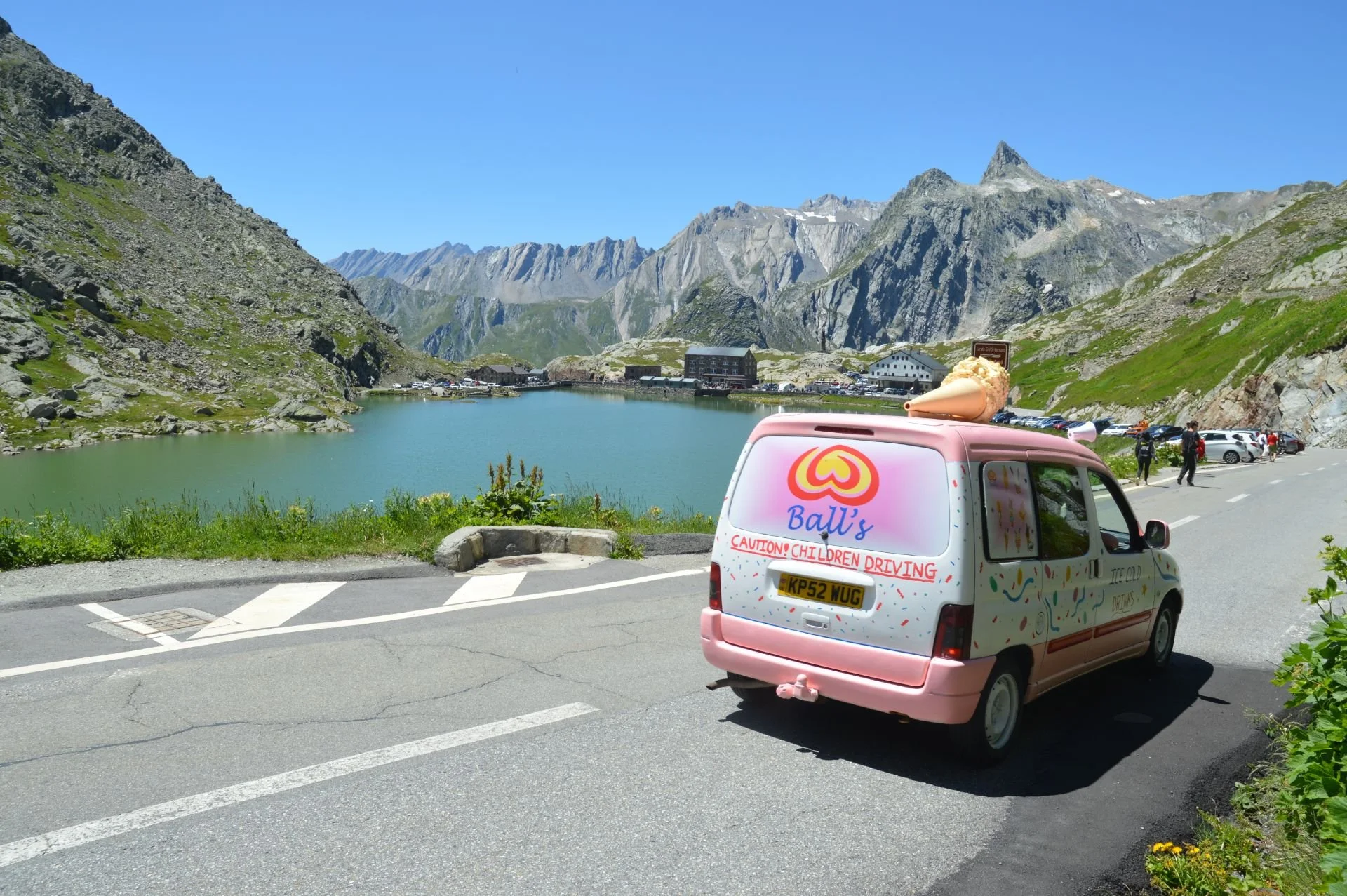 Pink ice cream truck with colorful decorations parked on a mountain road near a lake, mountains in the background, and a few people walking nearby.
