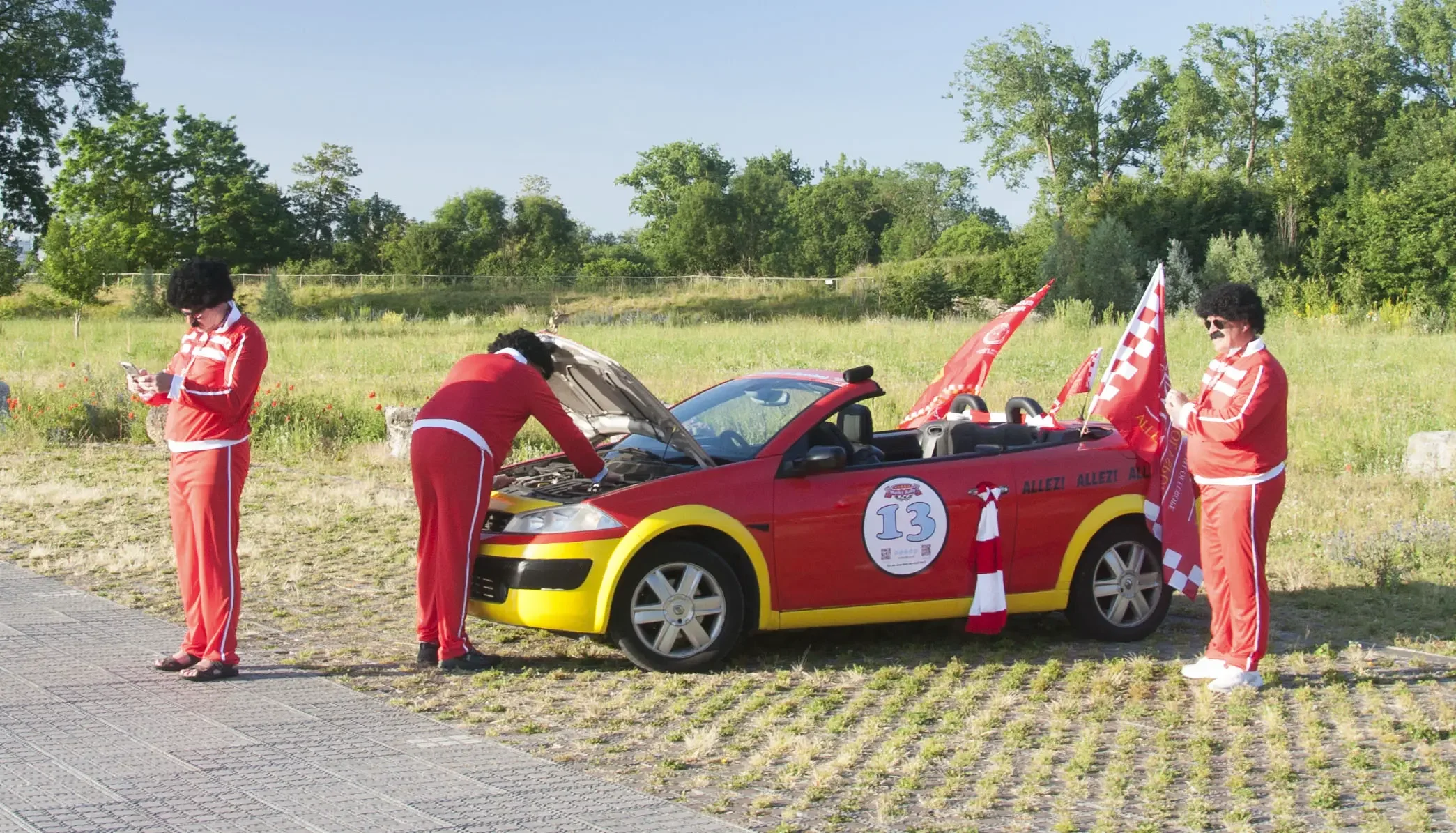 Three men dressed as Elvis Presley in red tracksuits with sunglasses and afro wigs are standing around a small red and yellow toy convertible car with racing flags, a large round number 13 sticker on the side, and a red and white checkered flag. One 