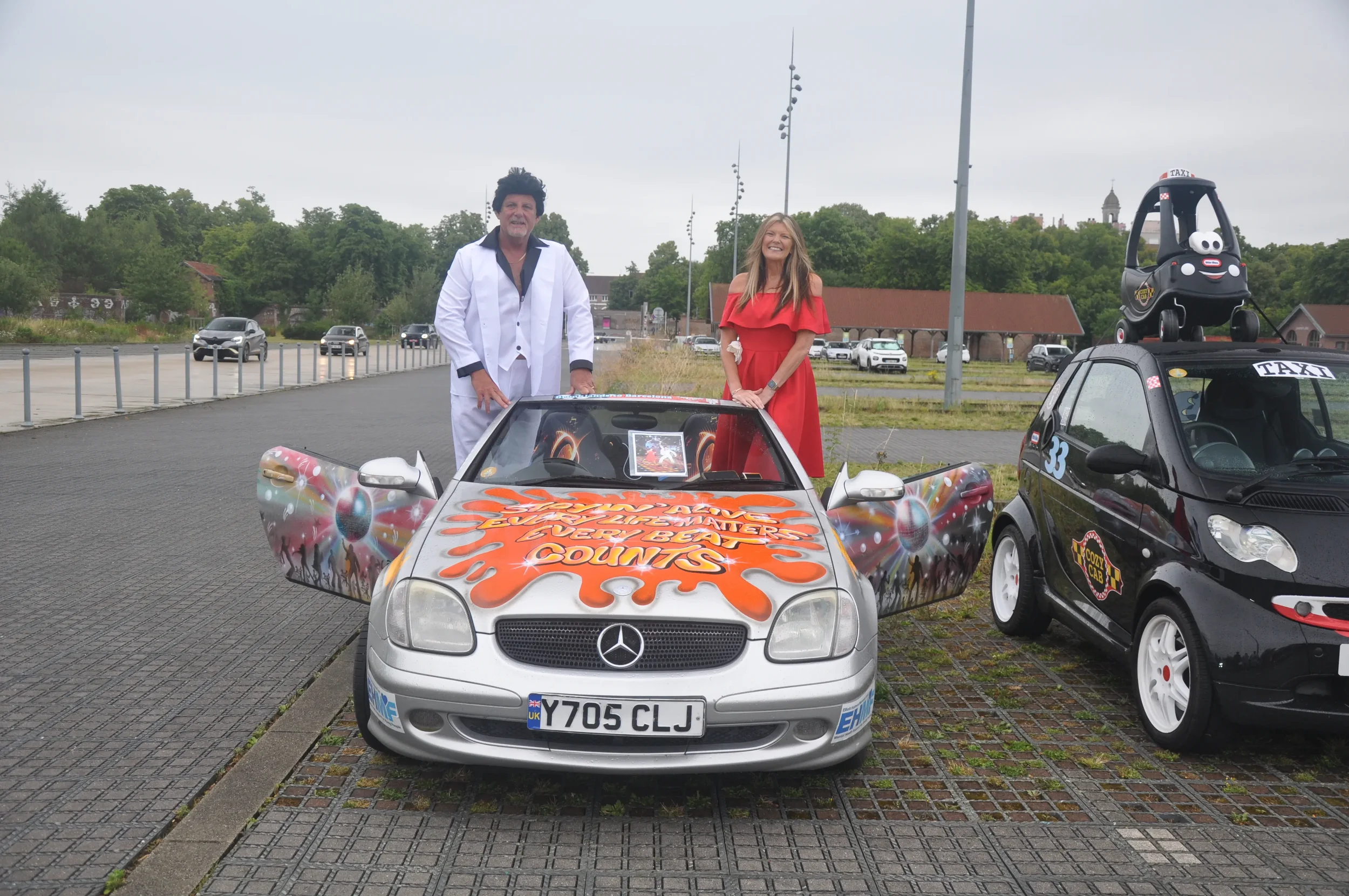 Two people stand beside a colorful, custom-painted Mercedes-Benz convertible car with the number plate 'Y705 CLJ.' A woman in a red dress and a man in a white suit are smiling at the camera. To the right, a small black car with a ride-on toy on top f